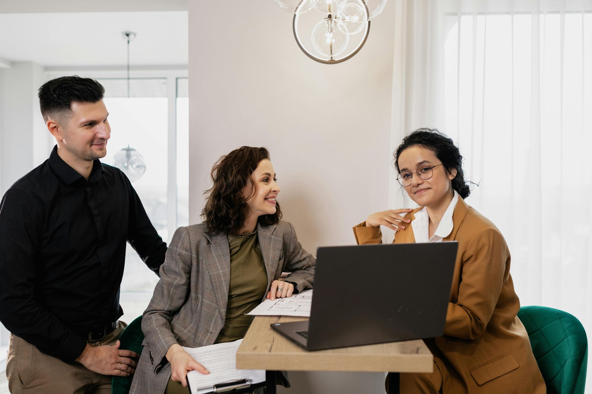 Three professionals collaborate around a laptop at a small table in a modern, bright office space.