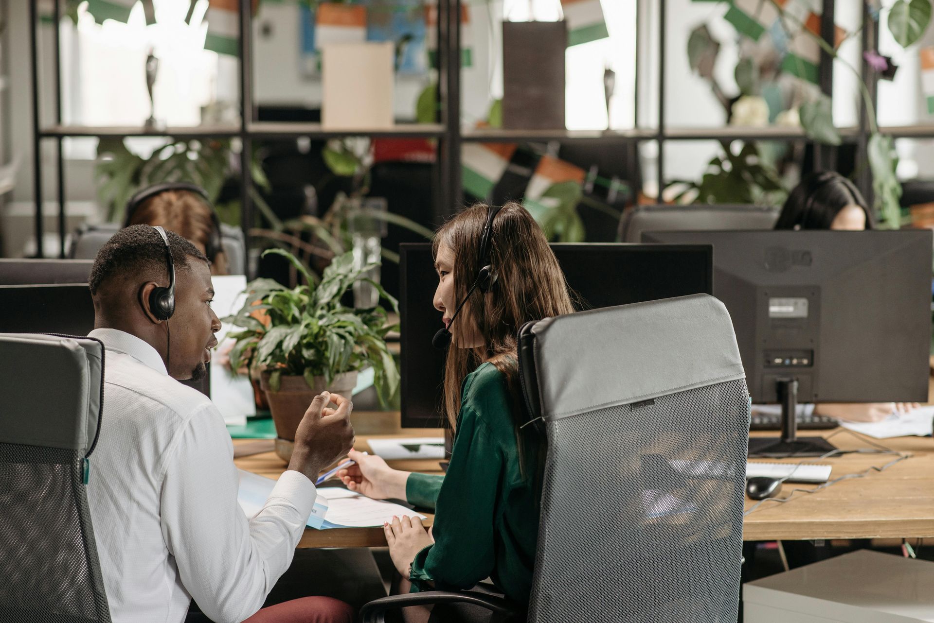 Two people with headsets at desks in an office; they're looking at papers, possibly collaborating.