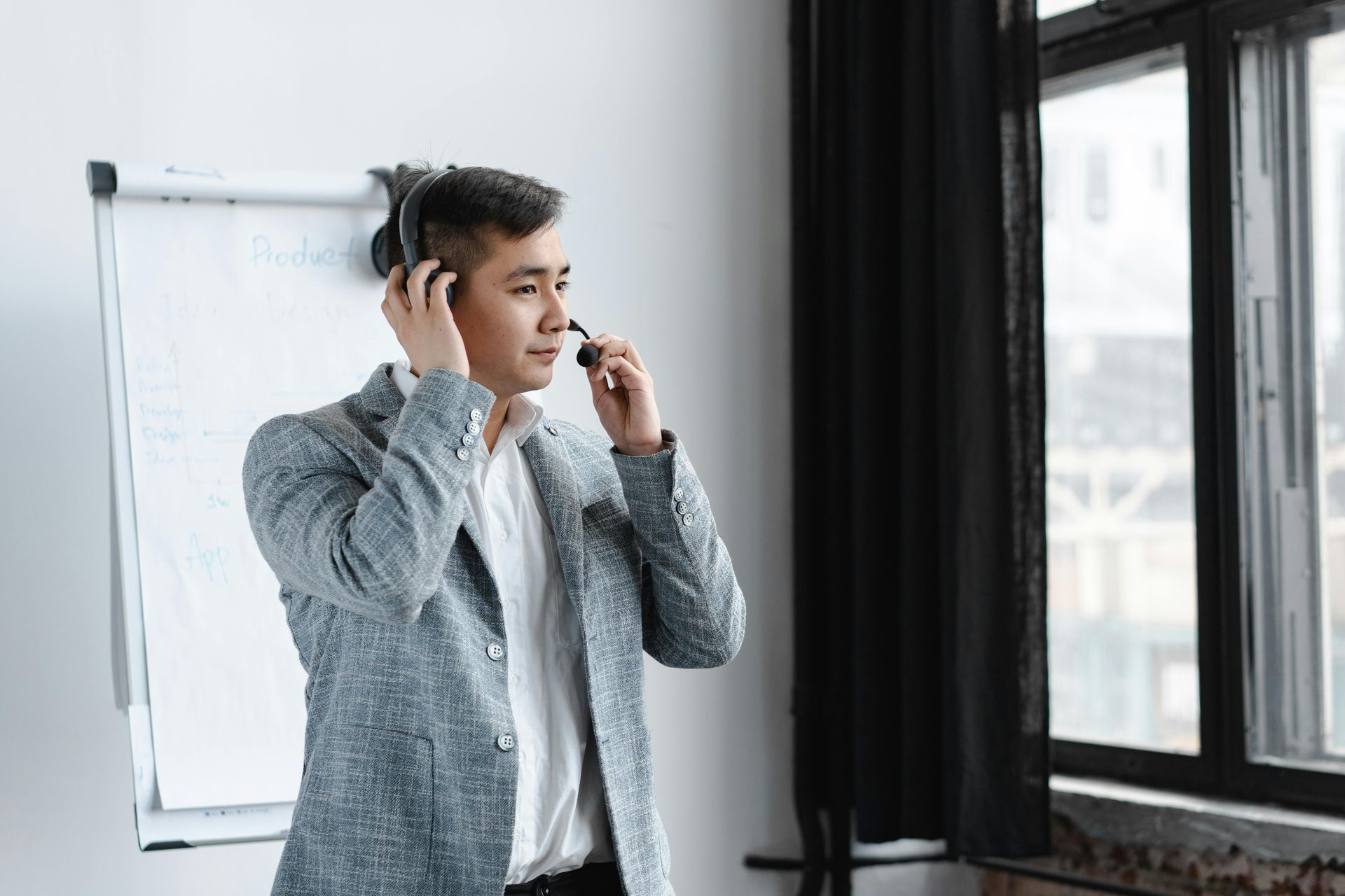 Man in a blazer adjusting a headset microphone near a whiteboard and window.