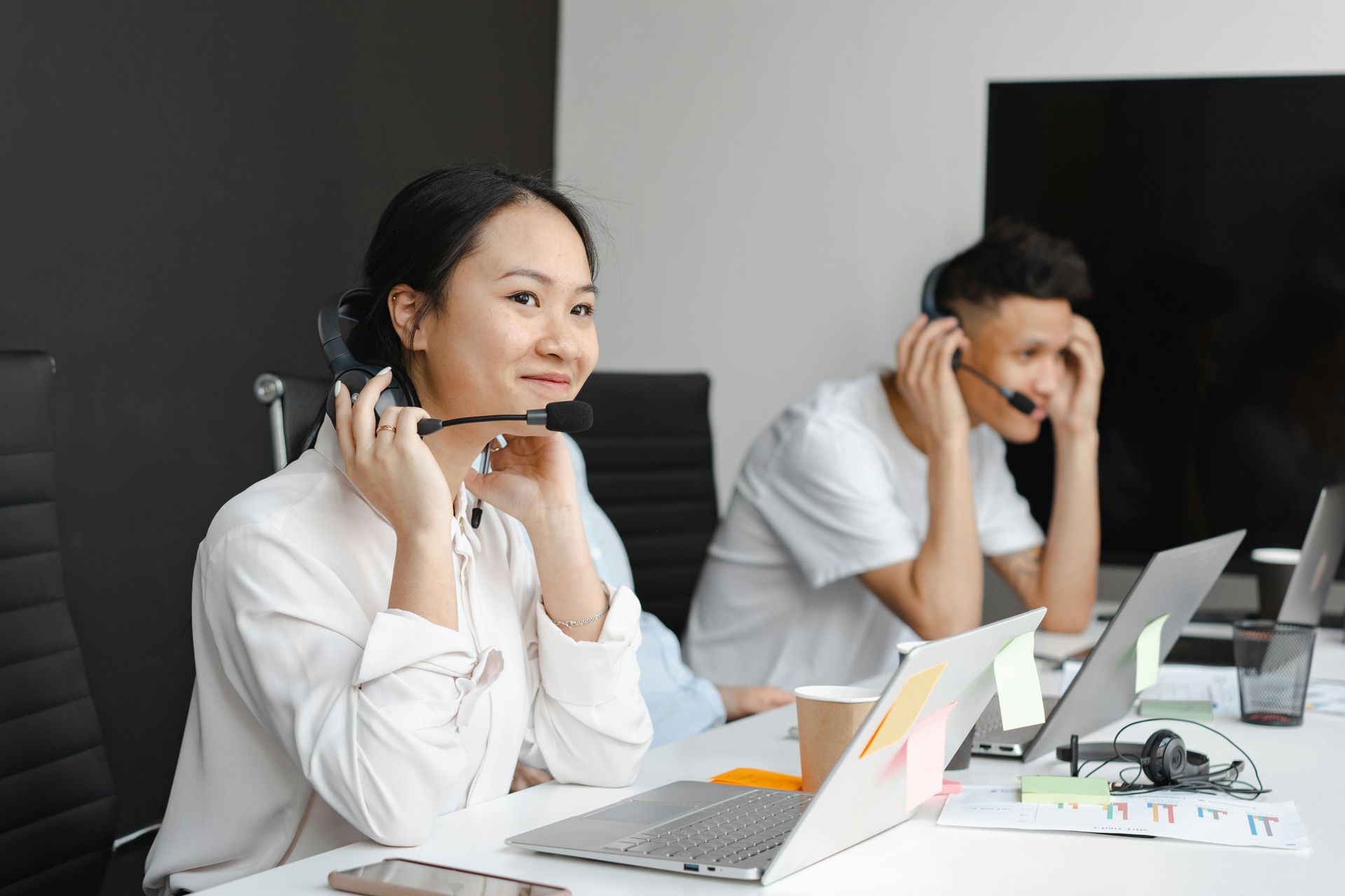 Woman and man in an office, wearing headsets, using laptops, and smiling.