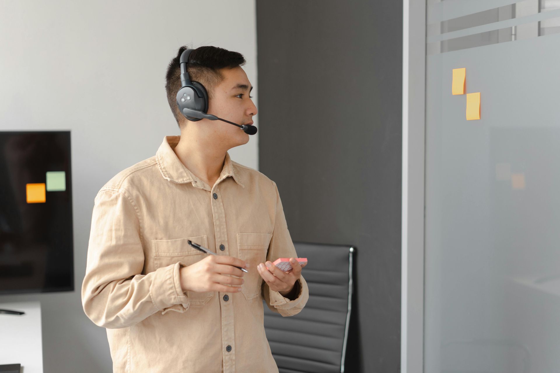 Man wearing headset, holding a pen, gestures while looking at a whiteboard with sticky notes in an office.