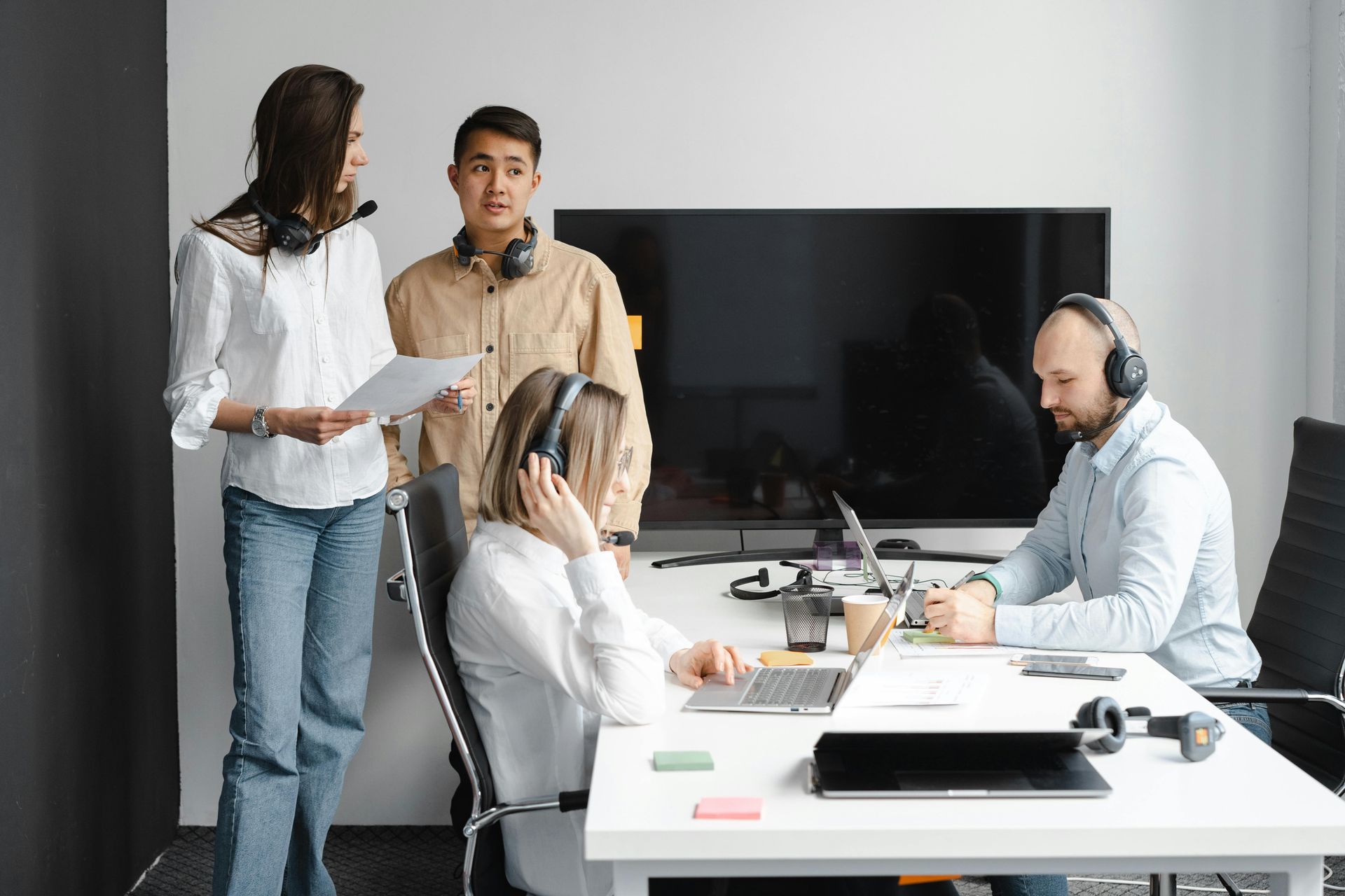 Four people wearing headsets in an office; two standing and two sitting at a table with laptops.