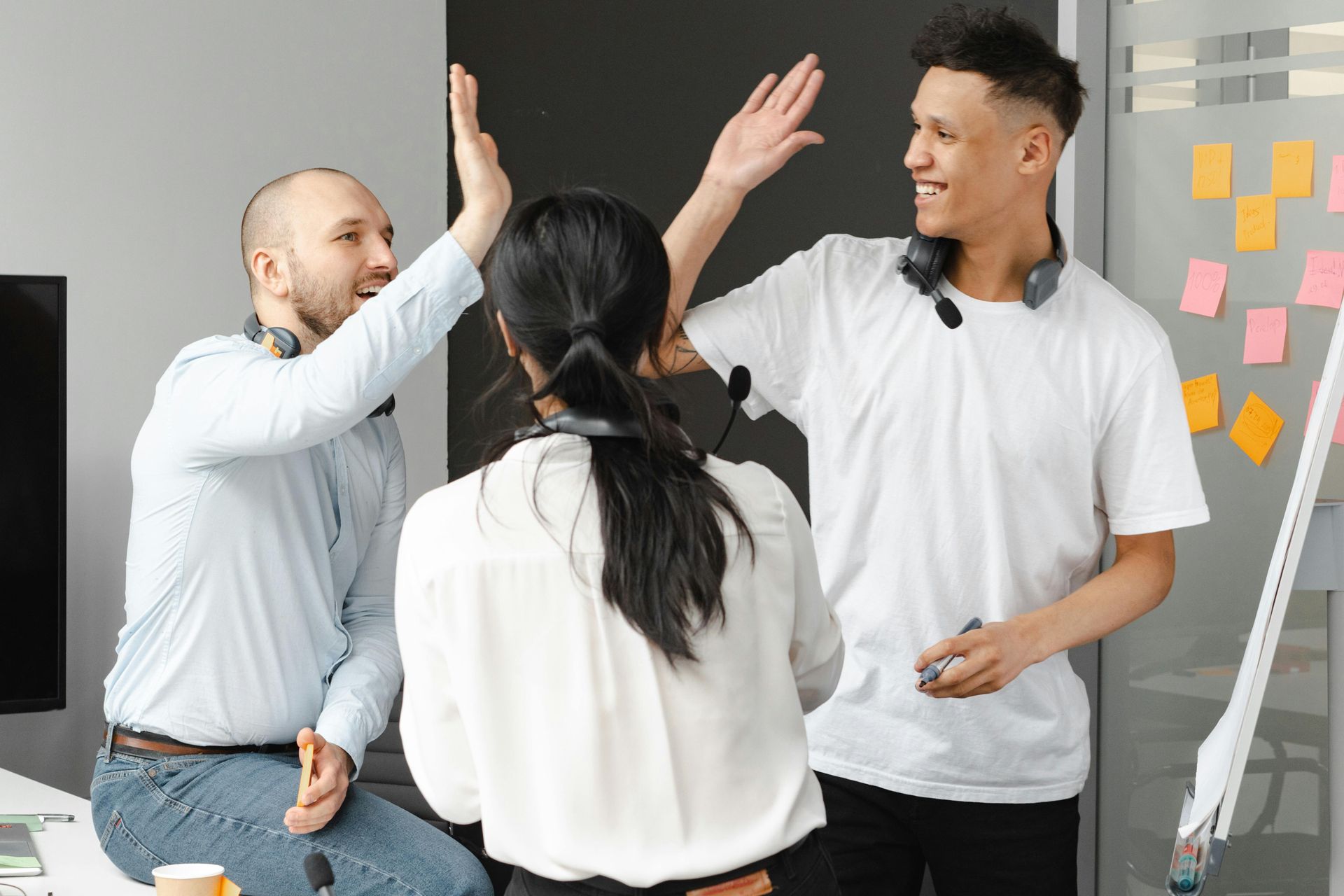 Three people in a modern office giving high fives. One person is sitting, all are smiling.