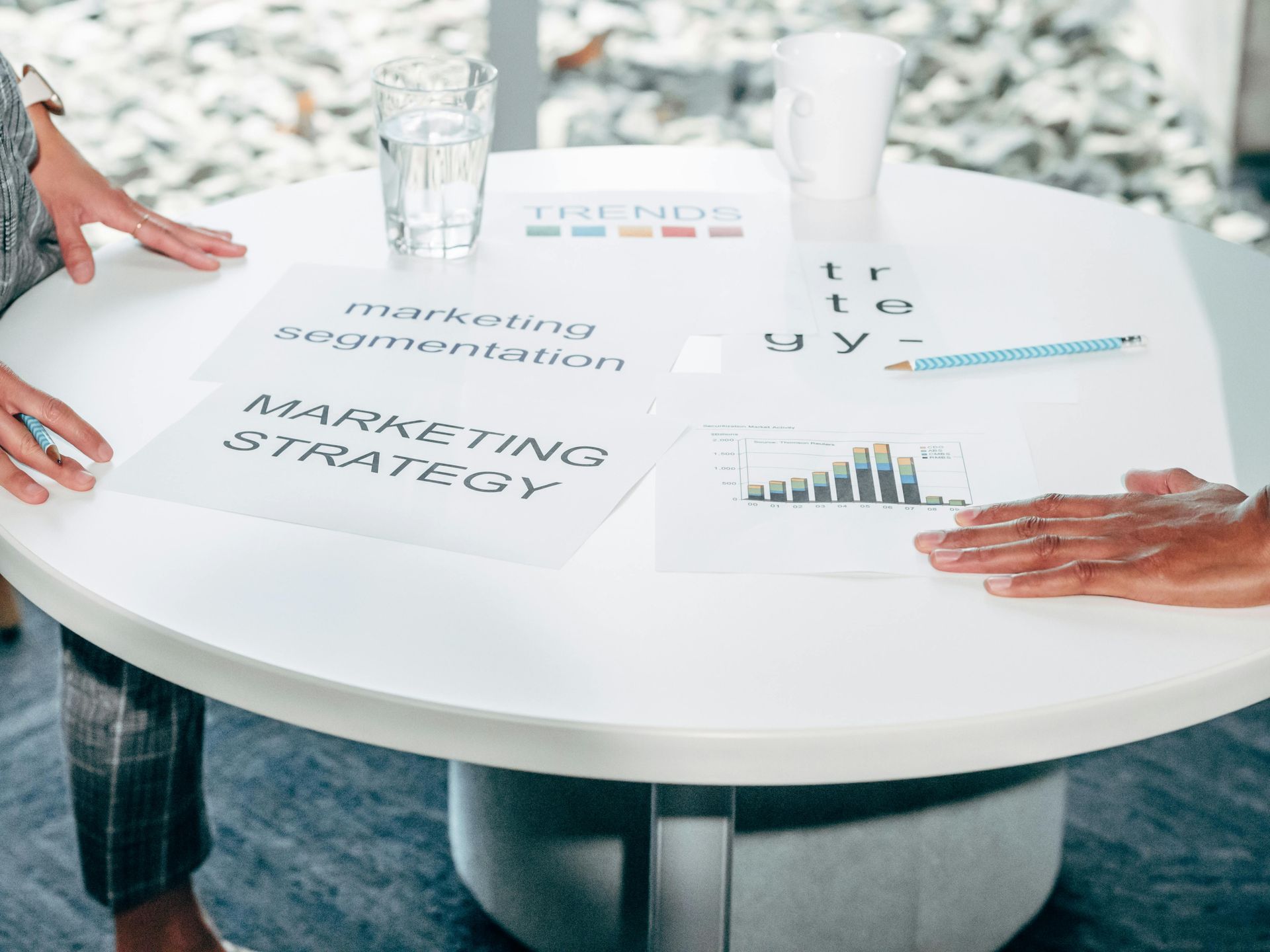 Two pairs of hands rest on a white round table featuring printed business strategy documents, charts, and a pen.