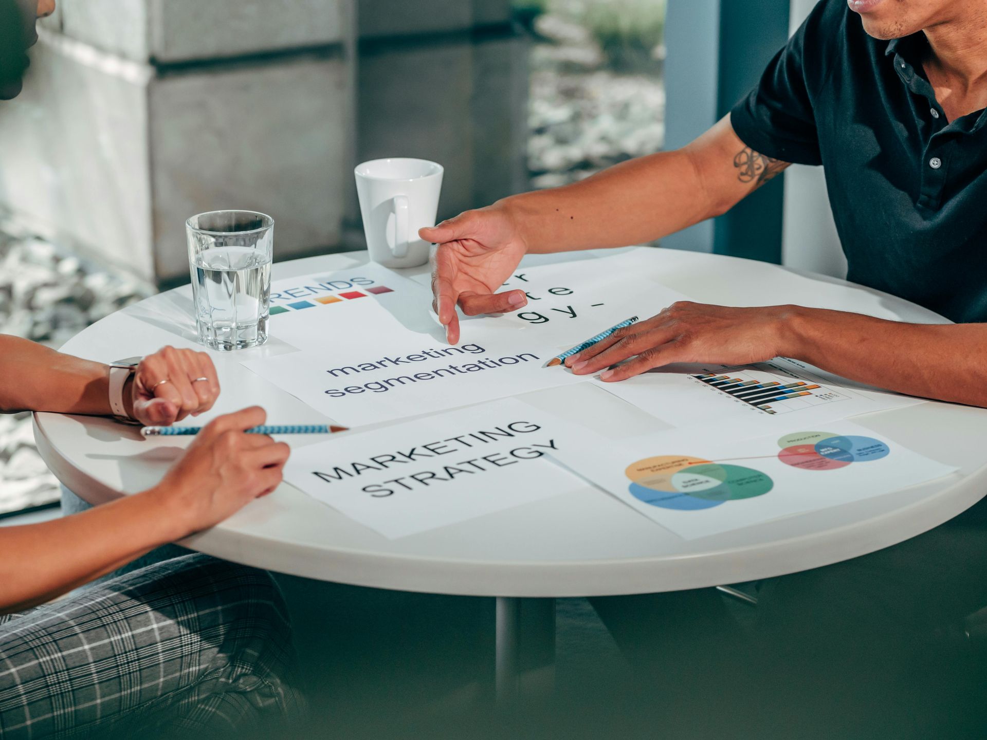 Two people sit at a round table, collaborating on handwritten notes and charts labeled 