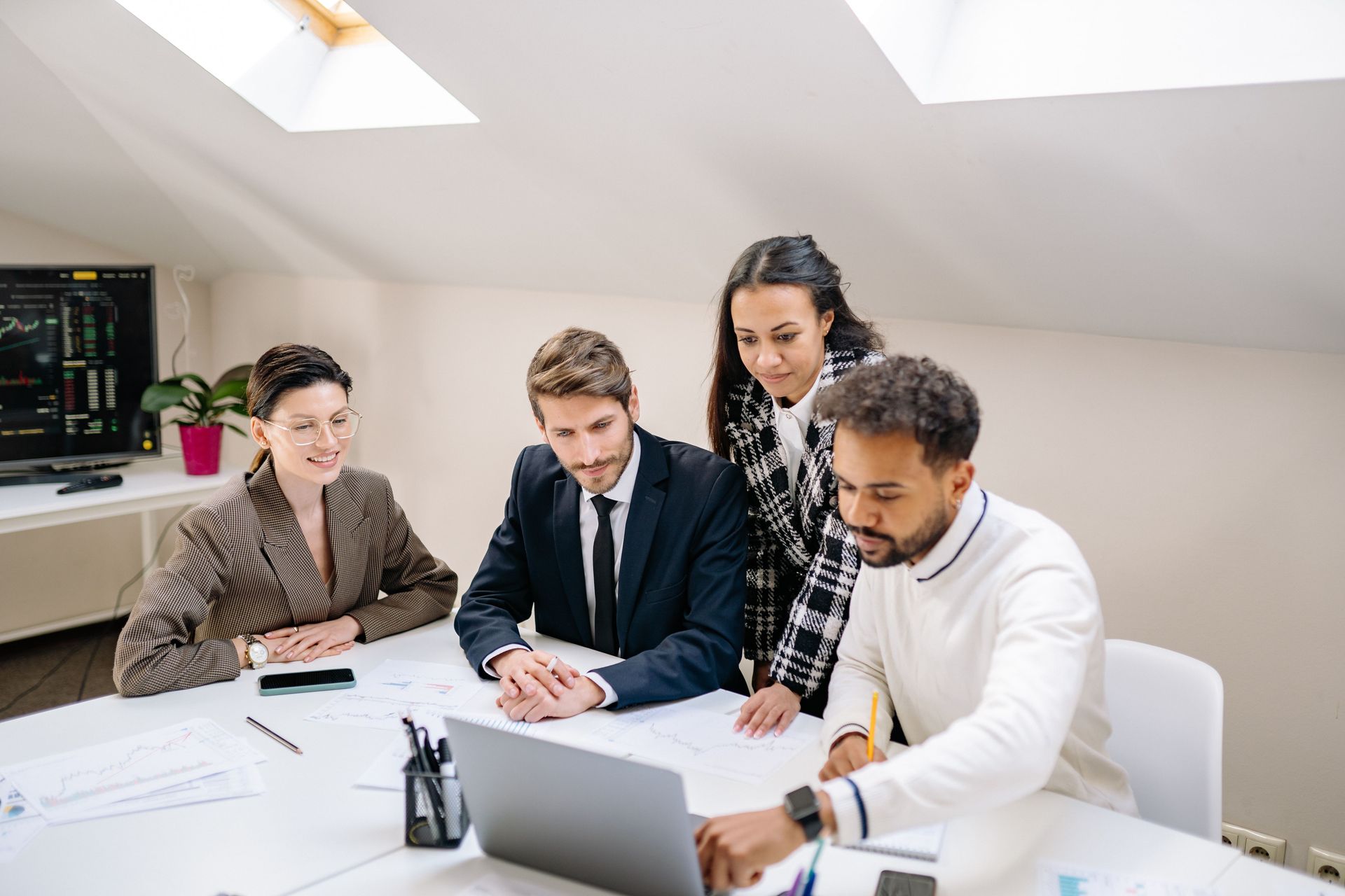 Four coworkers meet around a laptop and paperwork in a bright office conference room.