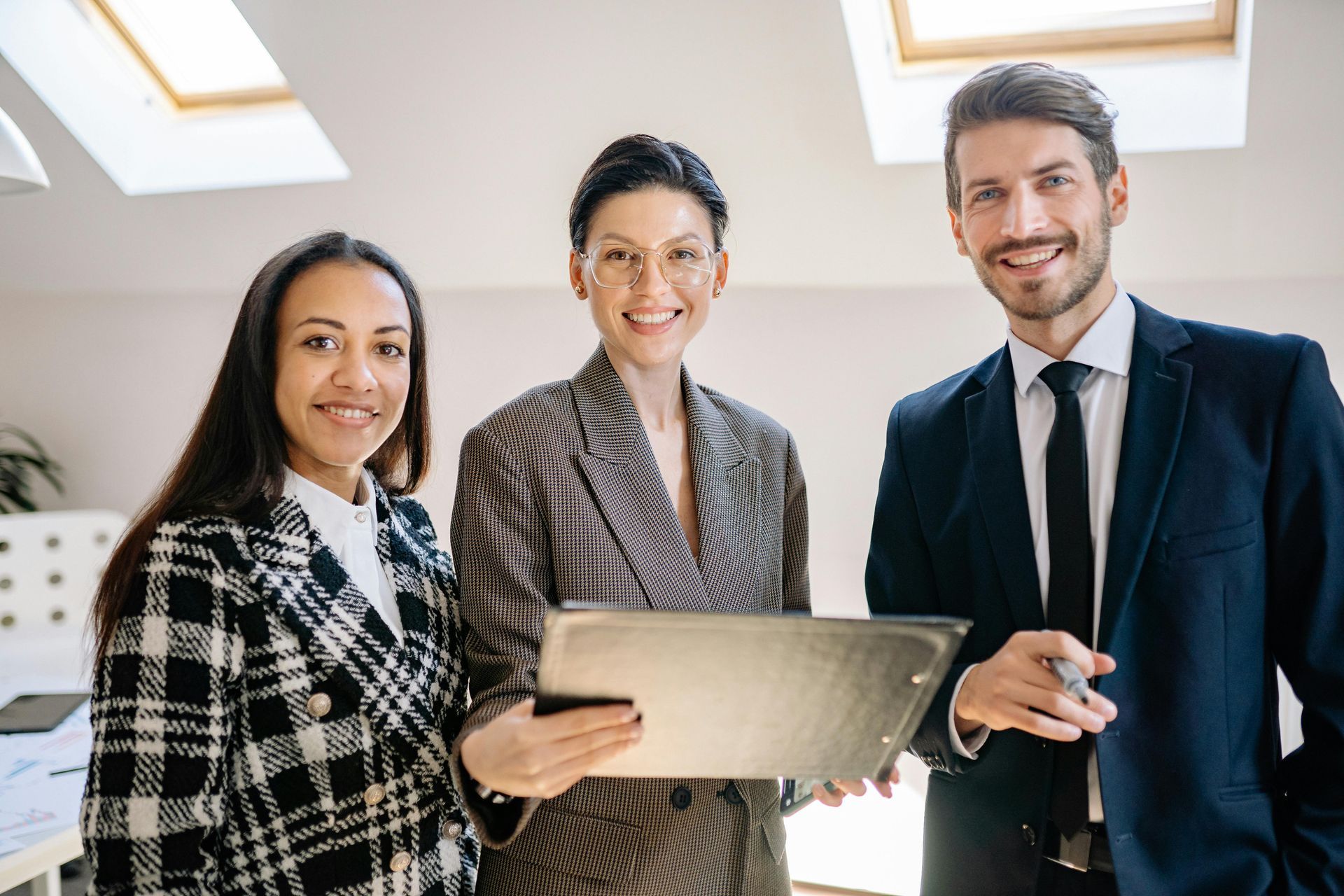 Three professionals in business attire stand together in an office, smiling as they review a document on a clipboard.