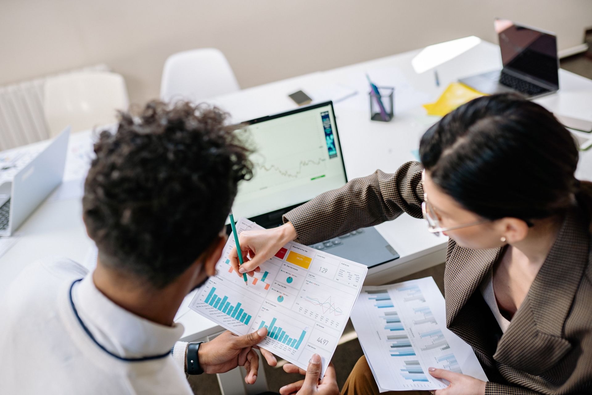 Two coworkers reviewing charts and notes at a laptop in a bright office meeting room