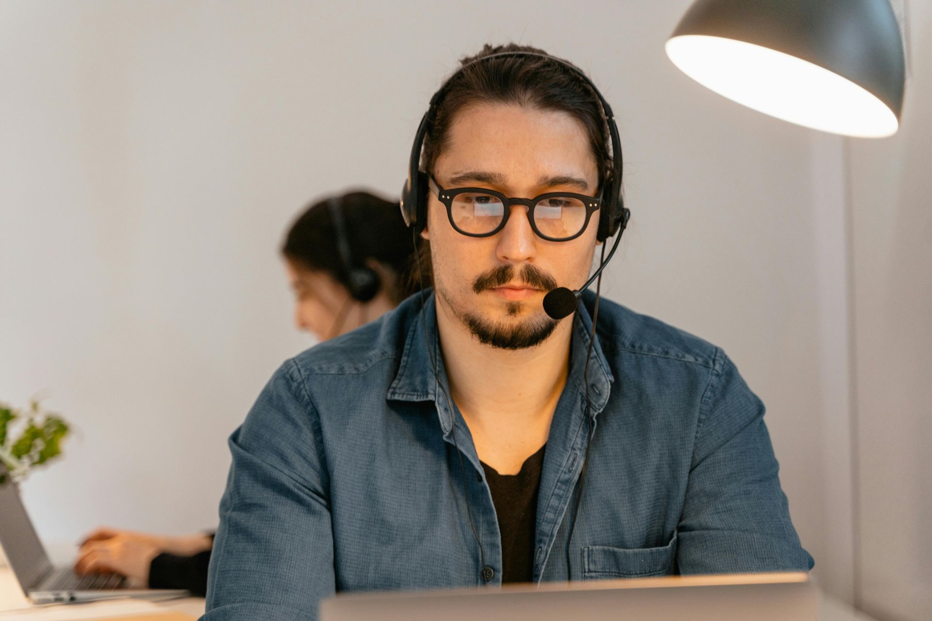 Man with glasses and headset at a desk, looking at the camera. Another person in the background also wears a headset.