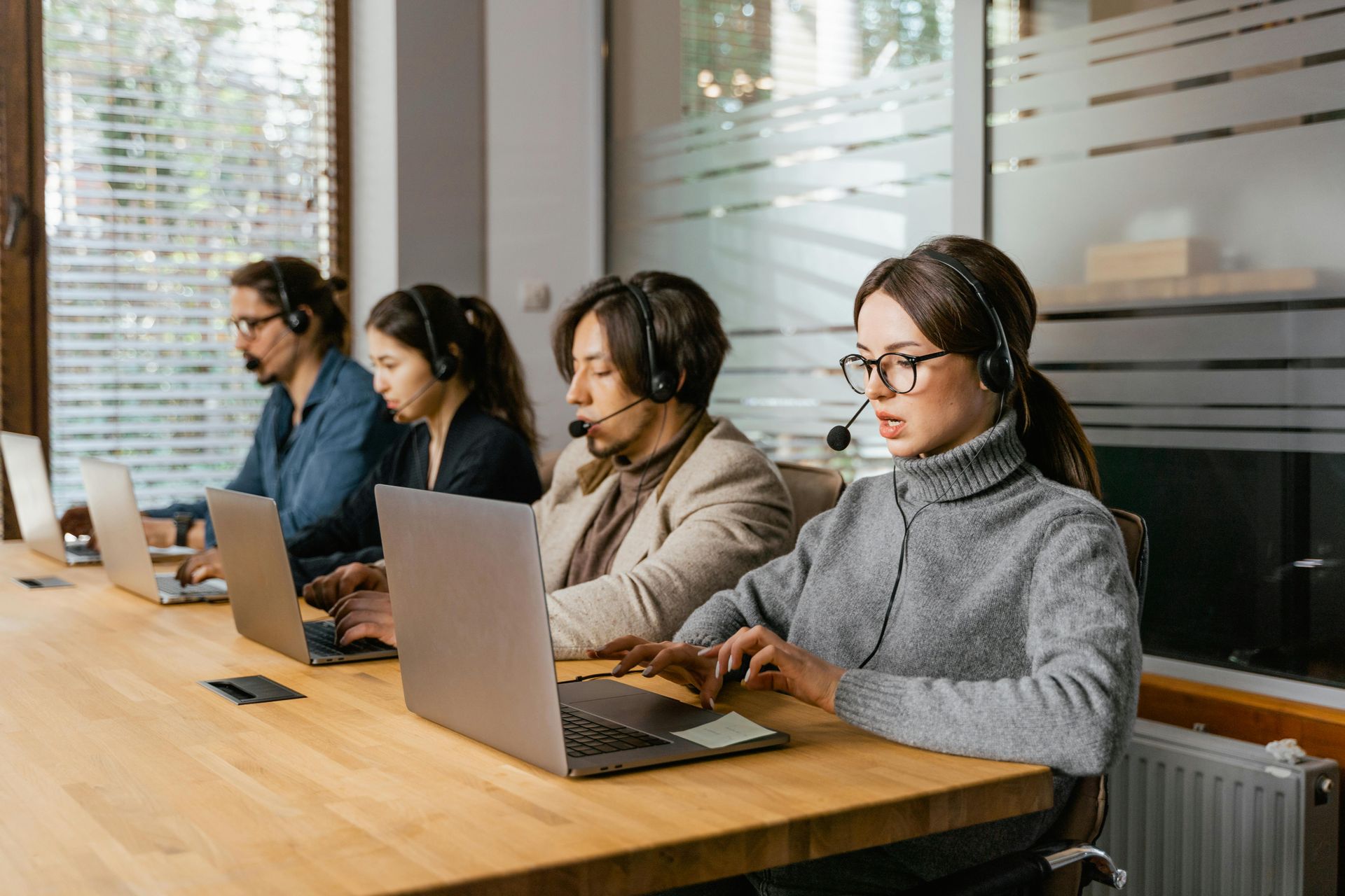 Four people with headsets sit at a long table, working on laptops. A modern office setting.