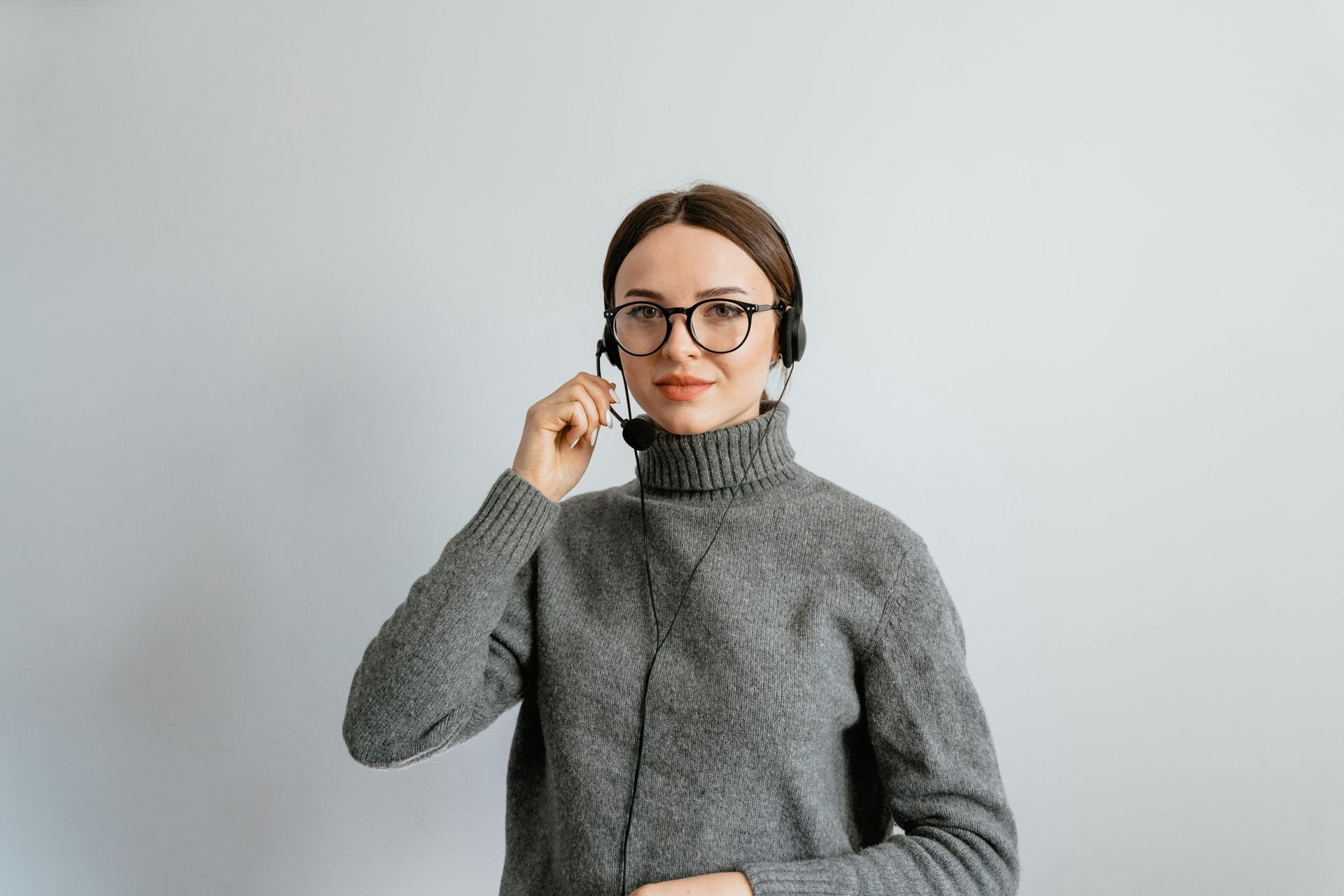 A person wearing a grey sweater, glasses, and a headset adjusts the microphone, positioned against a plain grey background.