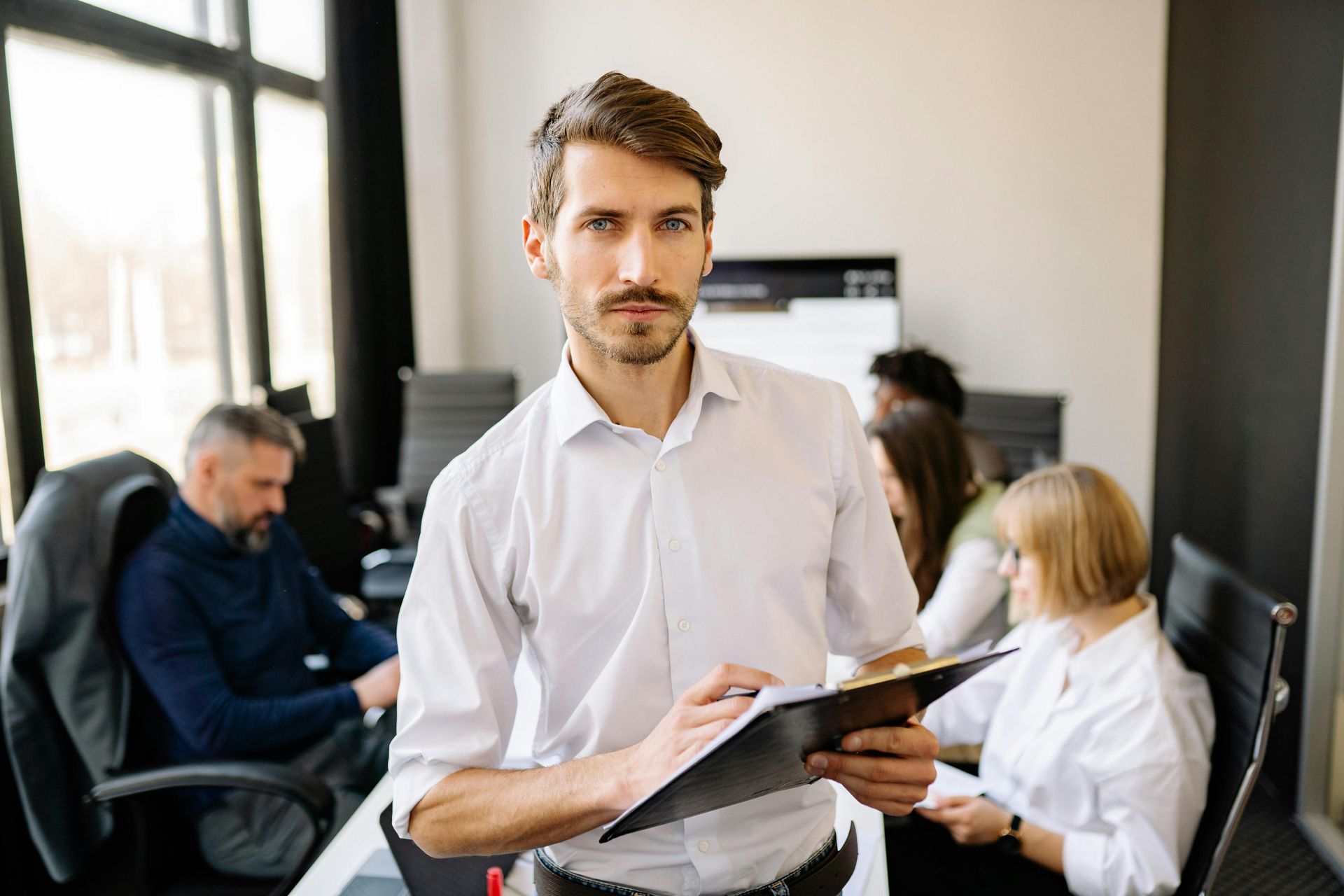 A professional man holds a clipboard in a bright office as colleagues work at desks in the background.