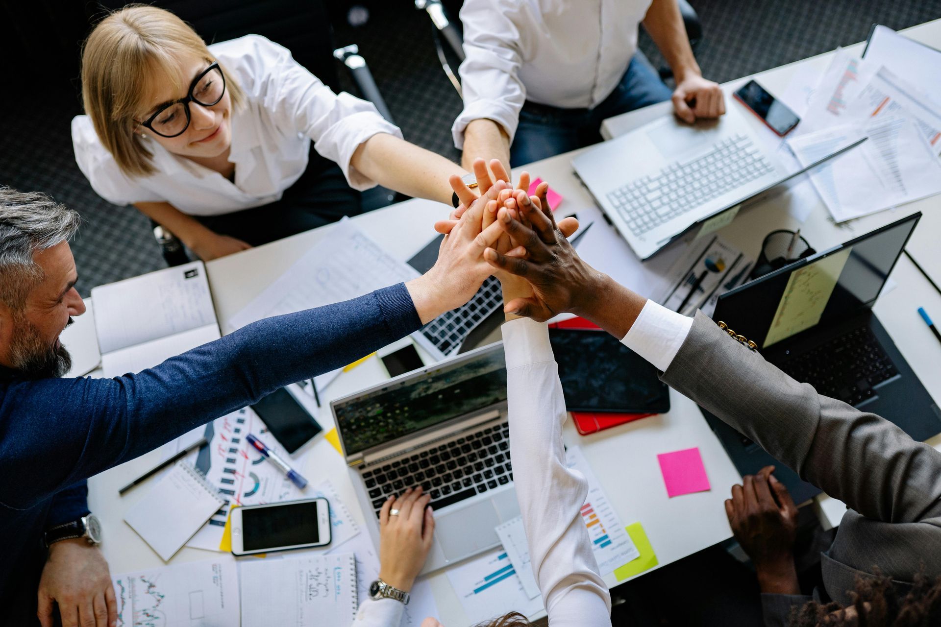 Colleagues gathered around a table stacking their hands together in a display of teamwork, surrounded by laptops and papers.
