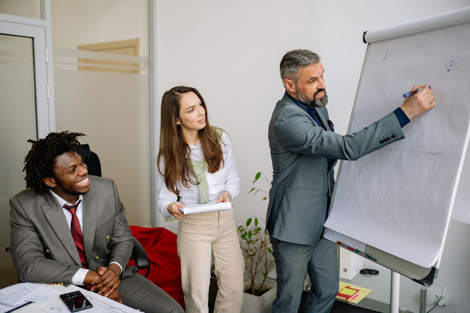 A man in a suit writes on a whiteboard while a colleague watches and another sits at a table in a modern office.