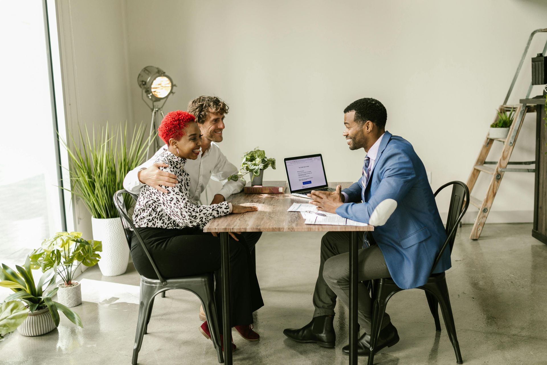 A person in a blue blazer speaks with a couple seated across a wooden desk in a modern, light-filled office.