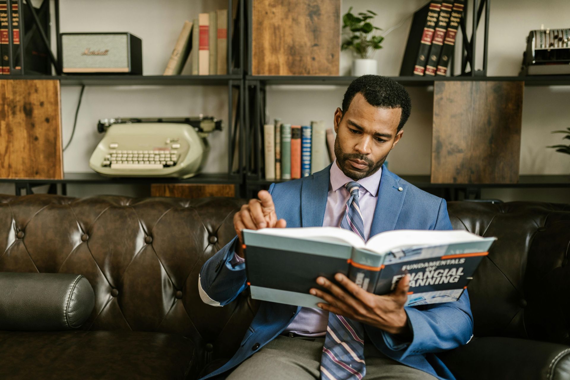 A person in a blue blazer and tie sitting on a leather sofa reading a financial planning book in a library setting.