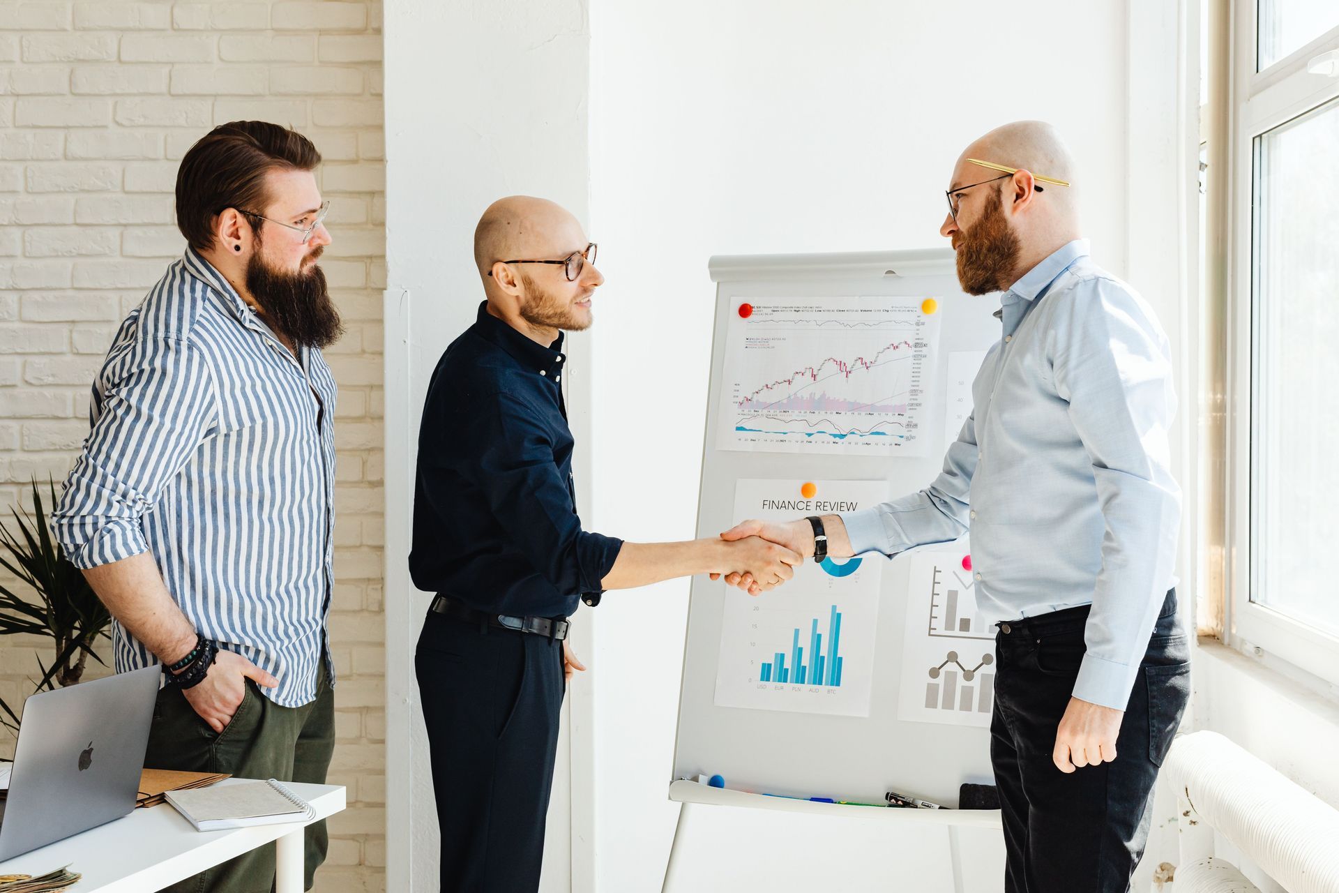 Three coworkers shaking hands beside a chart in a bright office meeting room