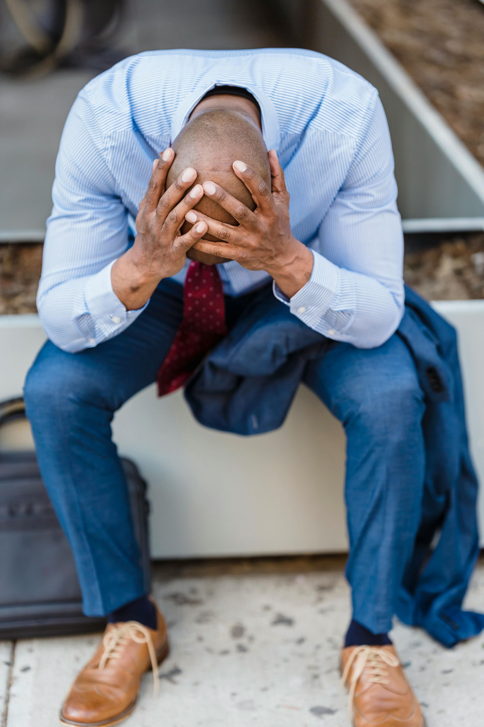 Man in business attire sitting with head in hands, looking distressed outdoors.