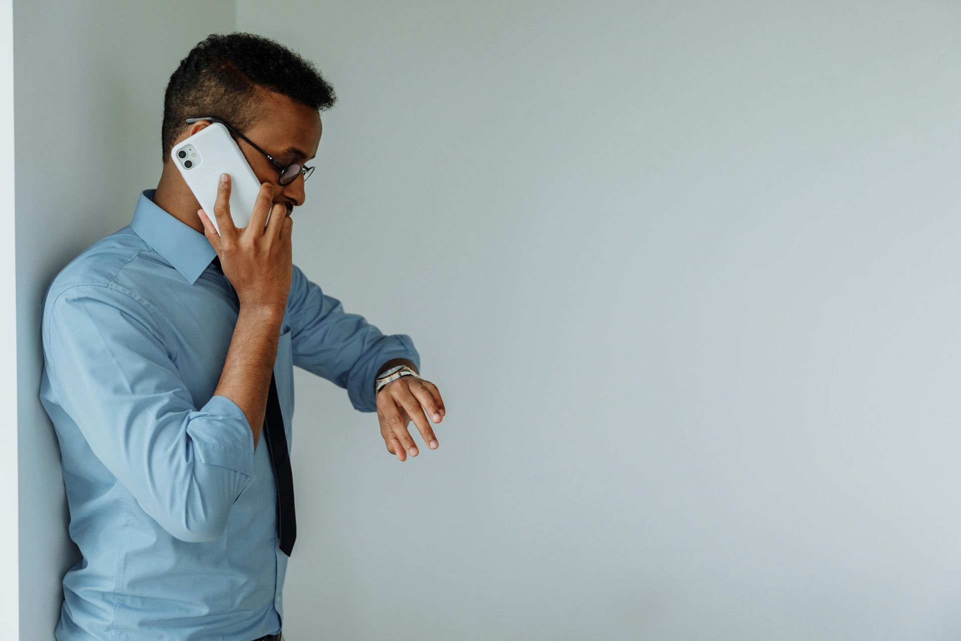 A person wearing a light blue dress shirt and tie leans against a wall while talking on a phone and checking a wristwatch.