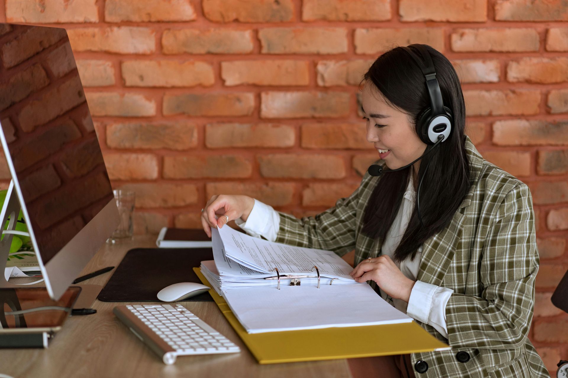 Woman with headset at desk, looking at documents. Brick wall background.