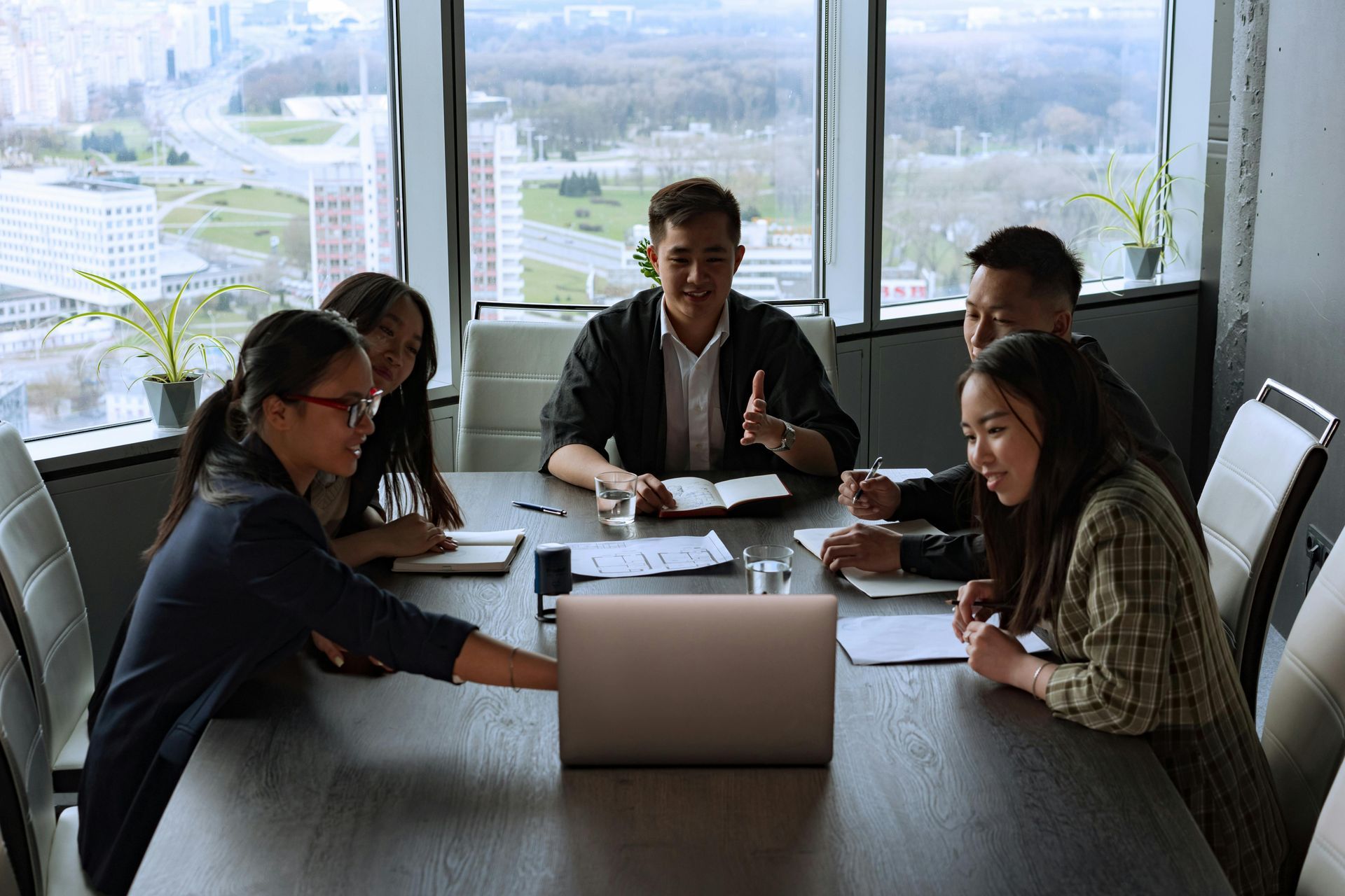 Five people in a meeting at a table, discussing work. A laptop and papers are on the table in a room with a city view.