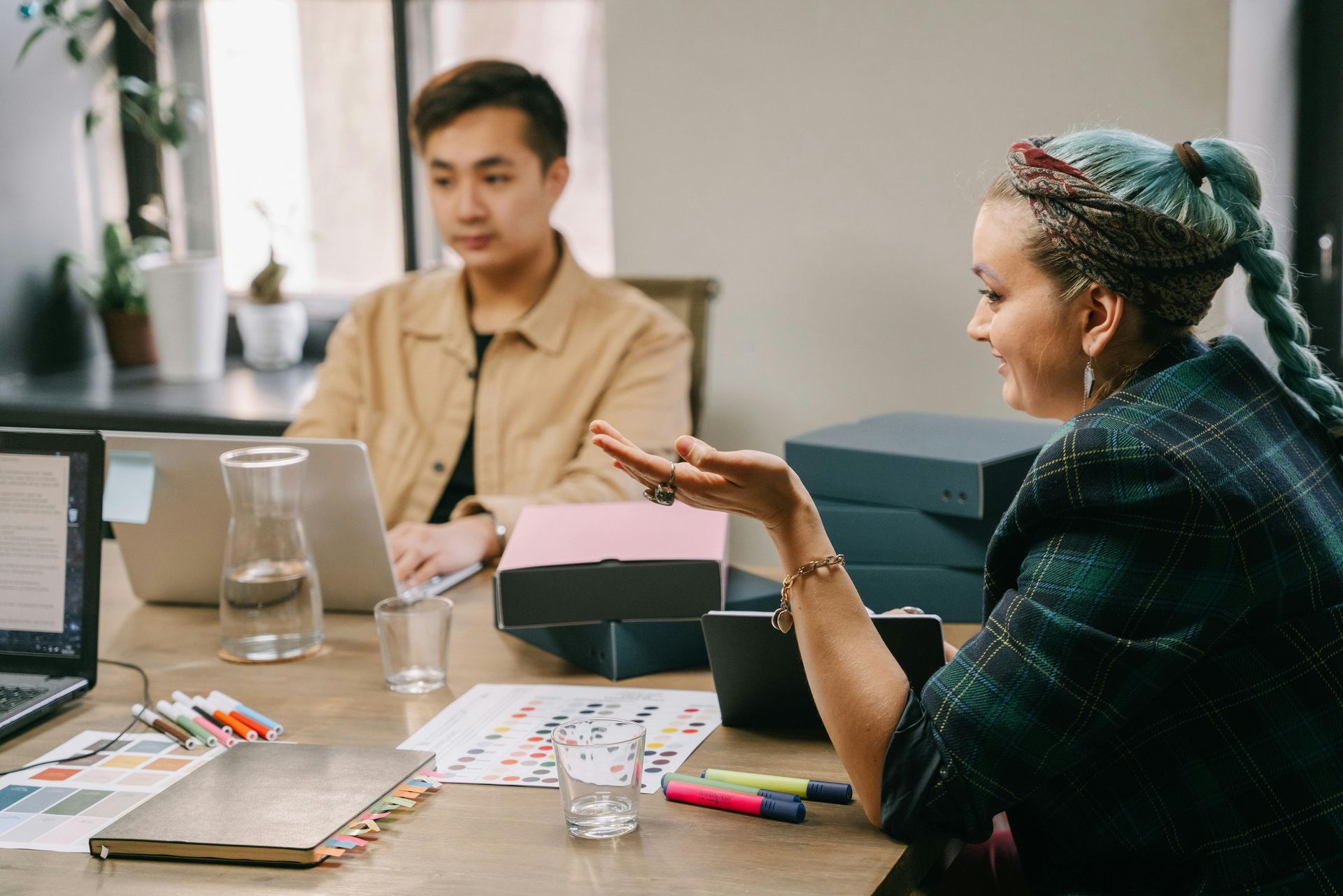 Woman with teal hair gestures while speaking to a person using a laptop at a table.