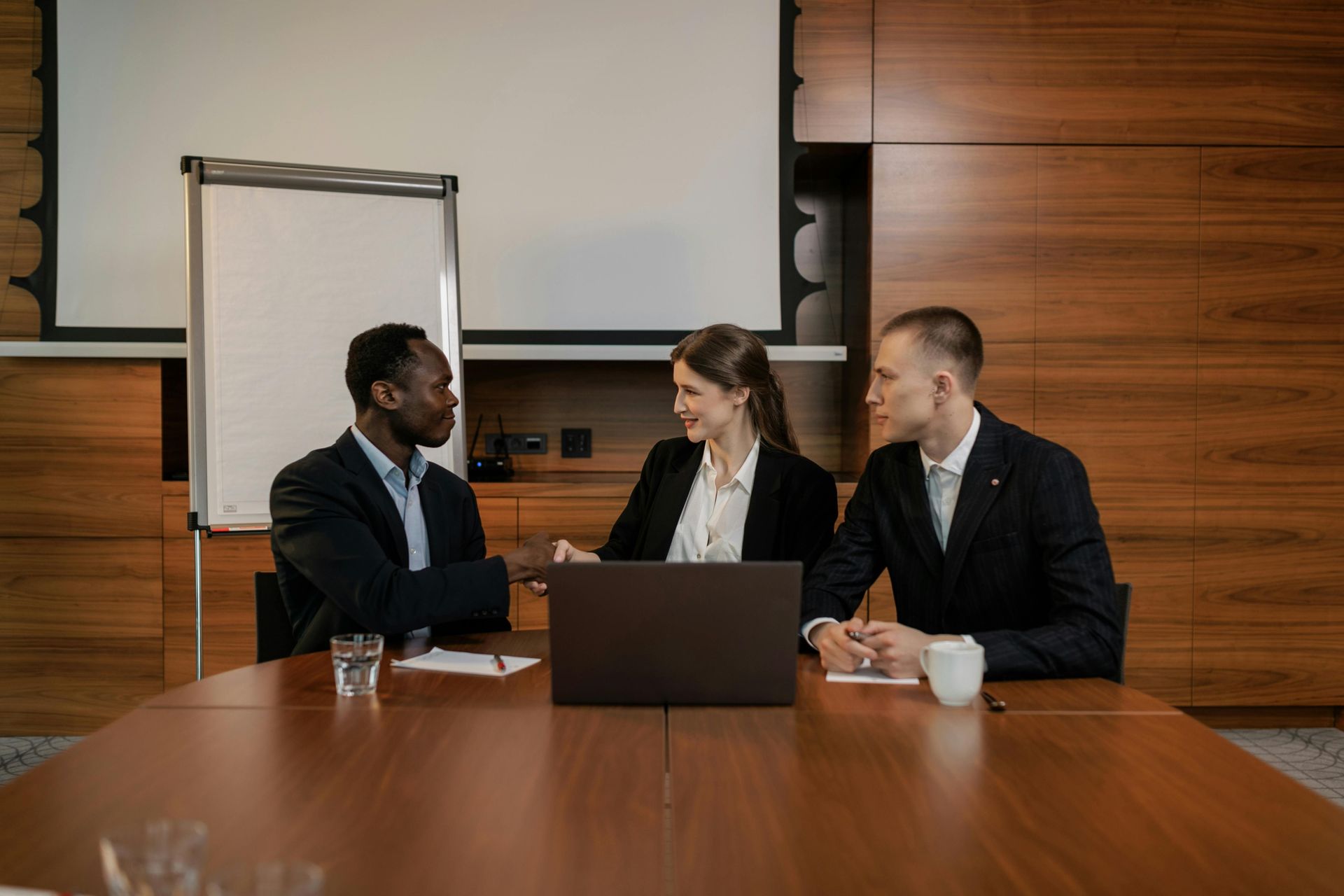 Three business people meet around a laptop in a conference room with wood-paneled walls.