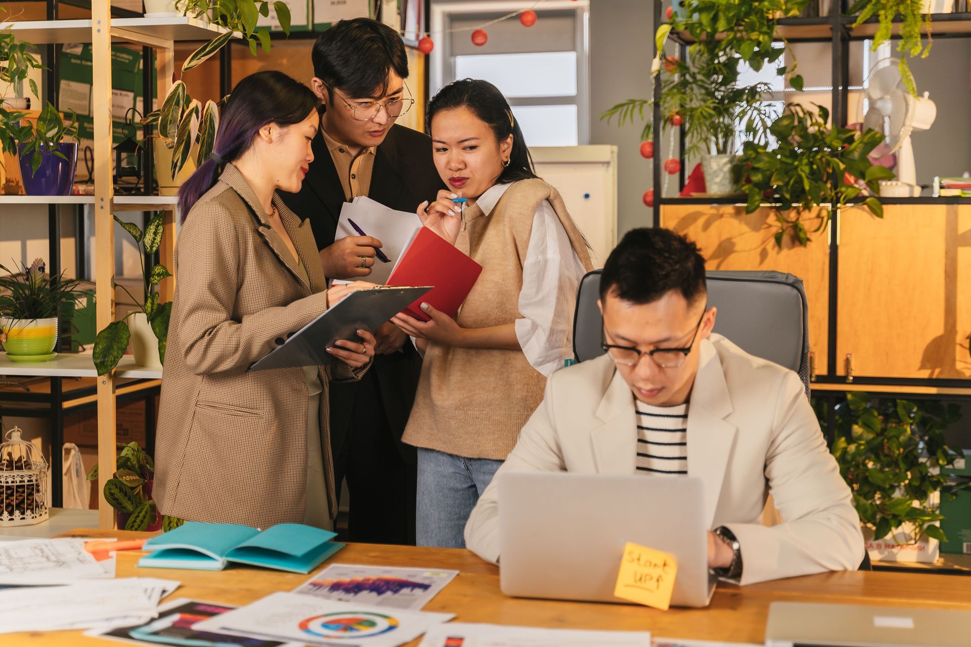 Four people collaborating in an office, gathered around a laptop and documents at a wooden table.