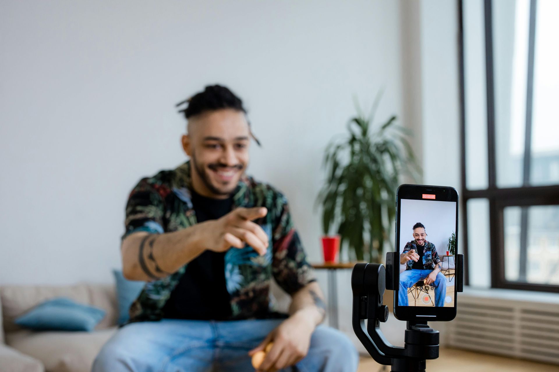 A person with braided hair sits on a couch, pointing at a smartphone mounted on a gimbal recording a video.
