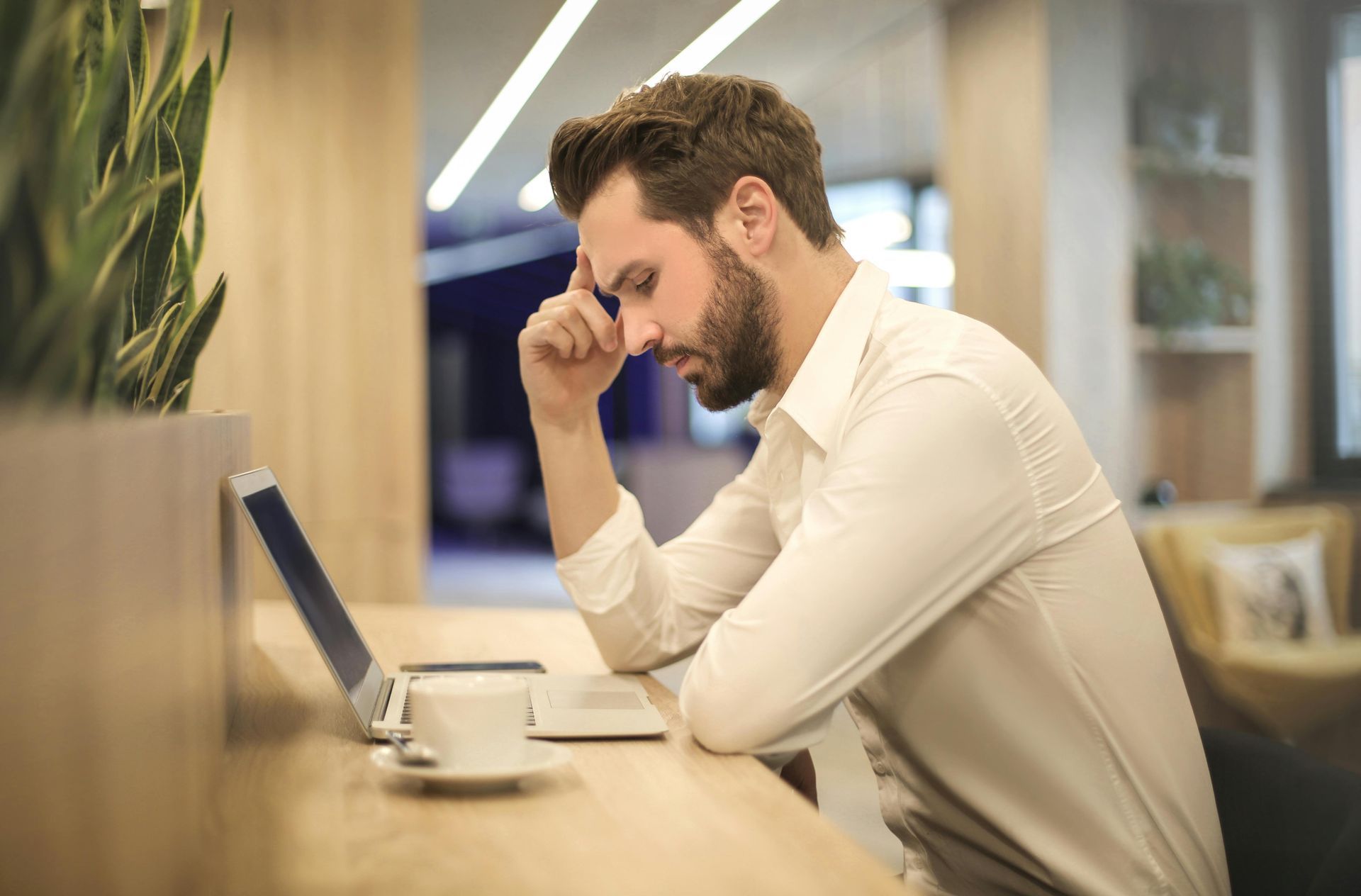 A person sits at a desk with a laptop and coffee, leaning their head on their hand while looking down, appearing stressed.