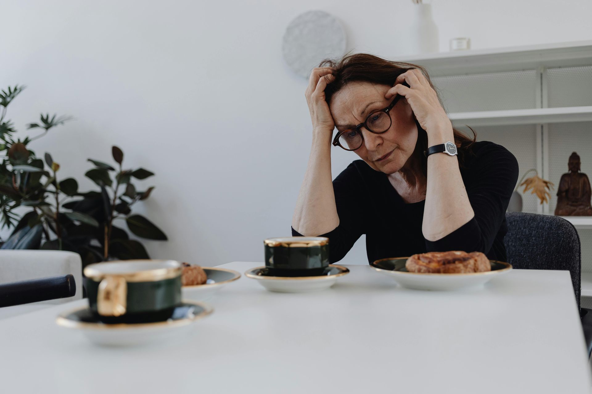 Woman with glasses holding her head at a table with coffee cups and pastries, looking stressed.
