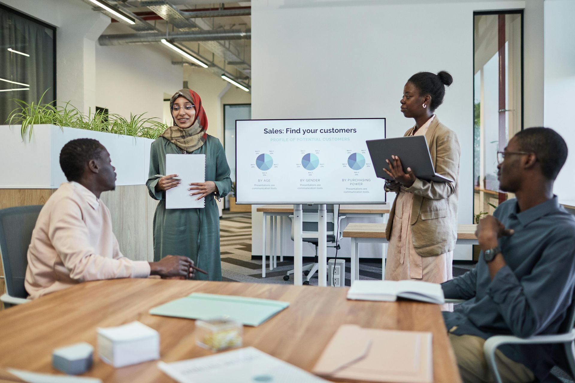 A group of colleagues in a bright, modern office meeting around a table to discuss a presentation on a large screen.