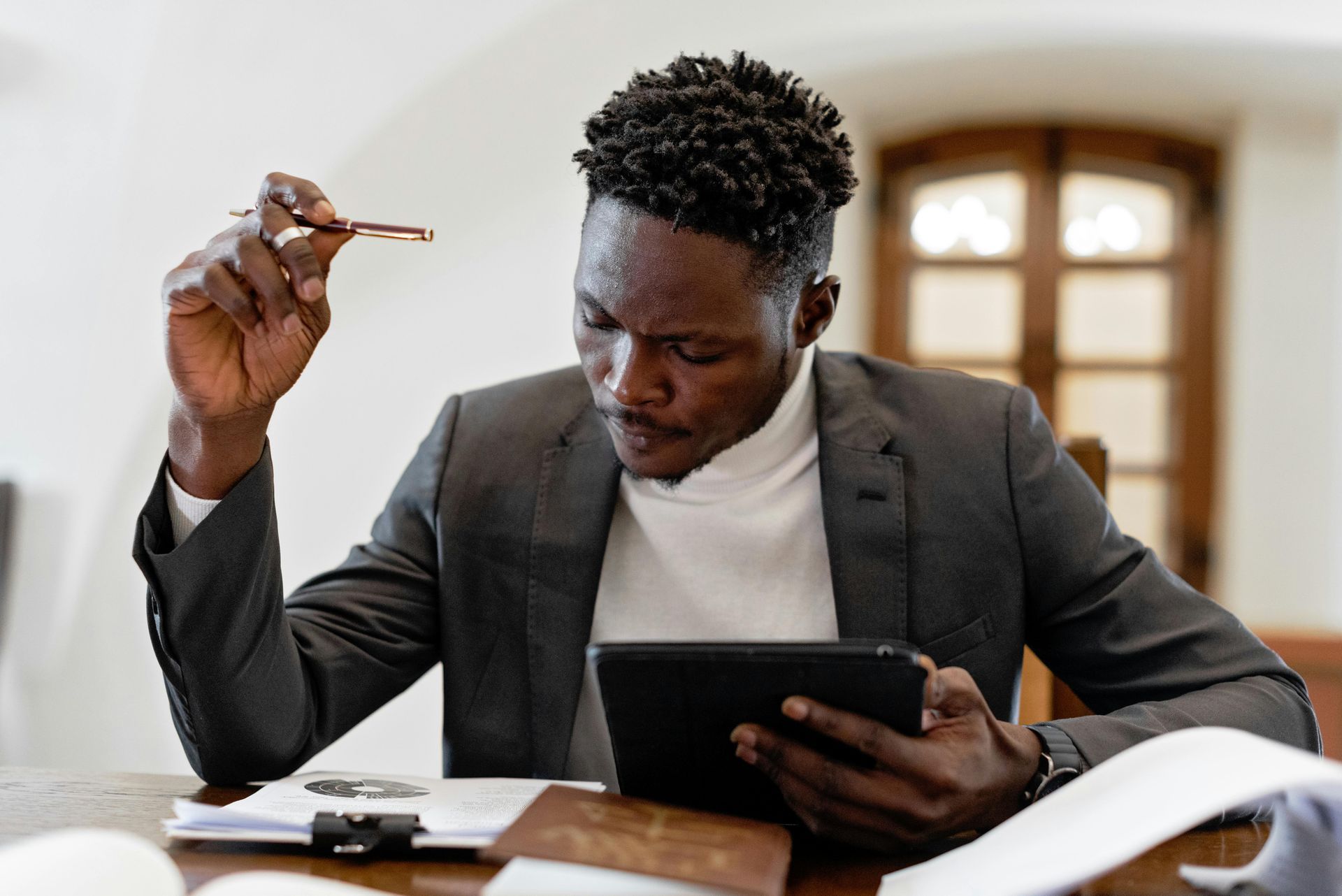 A professional in a grey blazer and white turtleneck reviews a tablet while sitting at a desk with paperwork.