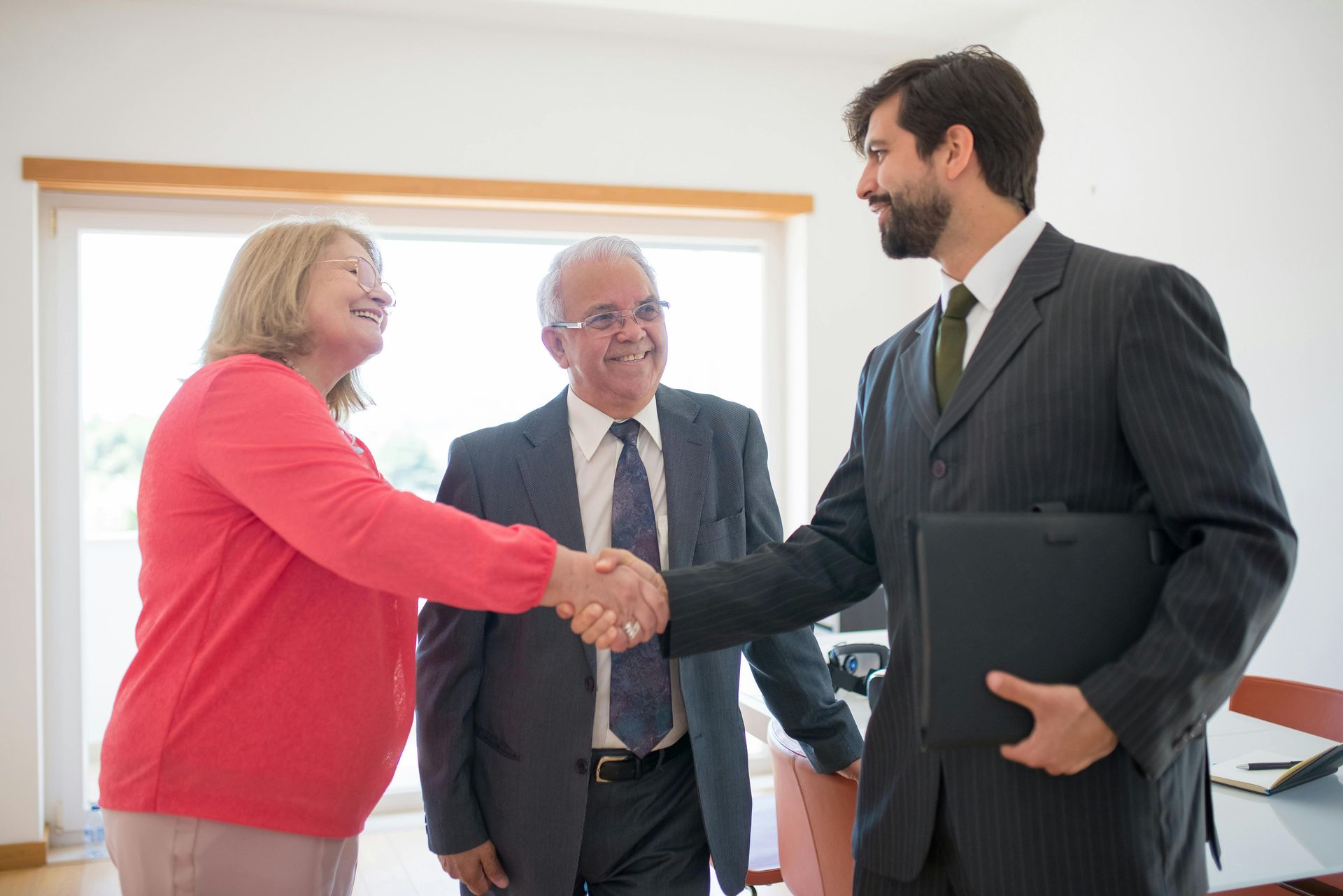 Man in suit shaking hands with woman, another man looks on; inside a room with windows.