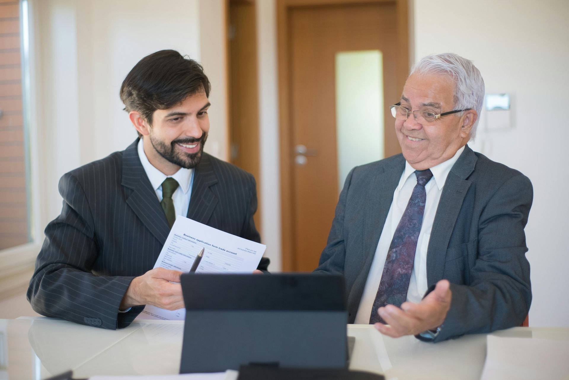 Two men in suits, one showing papers to the other, both smiling and sitting at a table with a tablet.