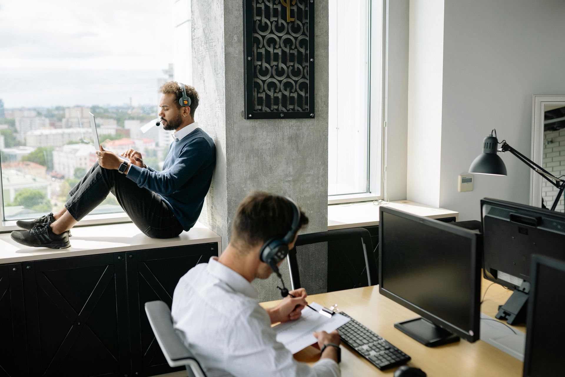 Two office workers with headsets work in a sunlit room; one sits on a window ledge, the other sits at a desk with a computer.