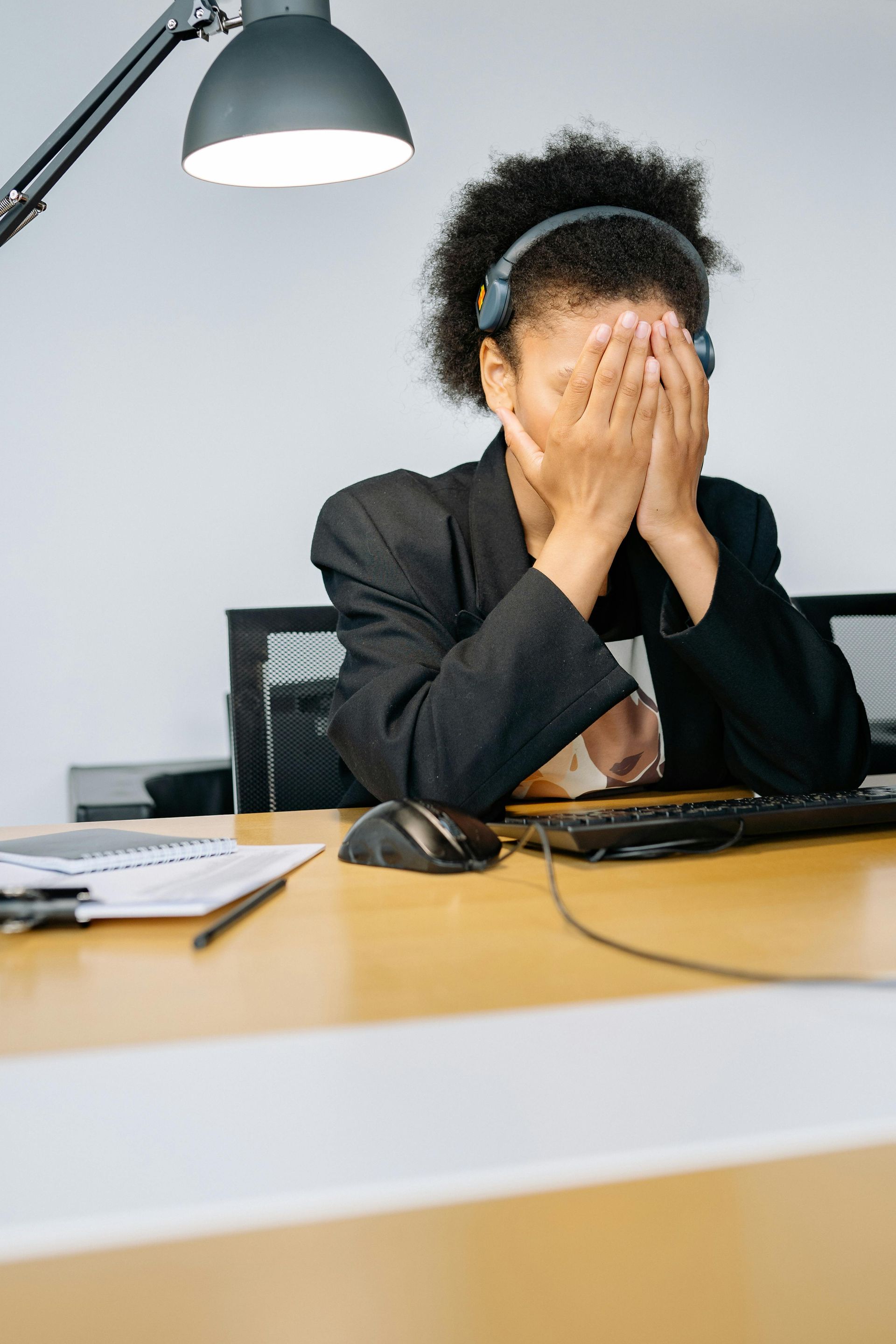 Person at desk covering face with hands; wearing headset and suit jacket.