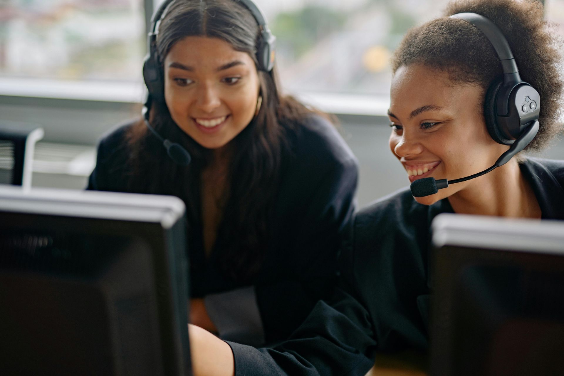 Two women in headsets smile while looking at computer screens in an office setting.