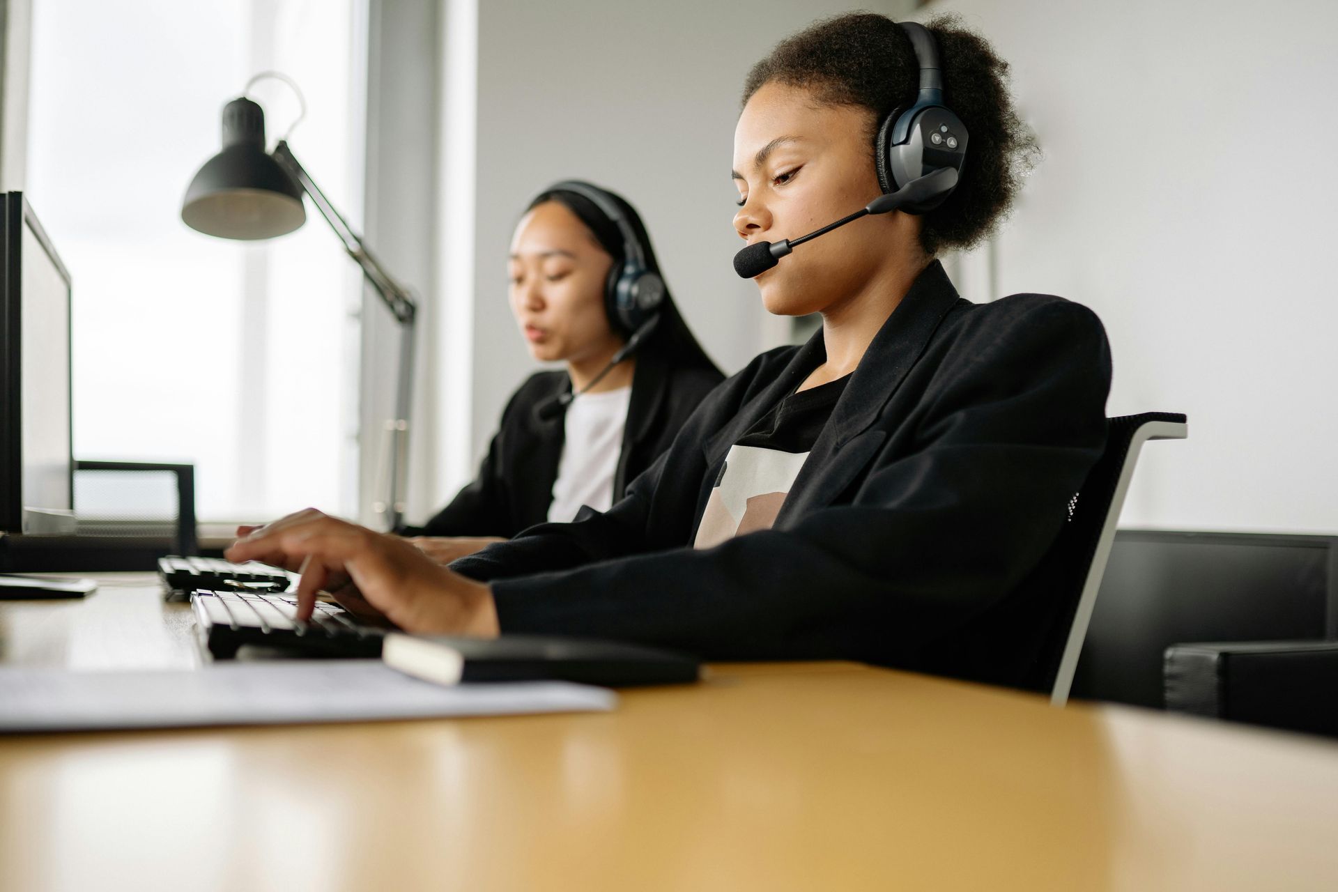 Two professionals wear headsets and work at computers in a bright, modern office.