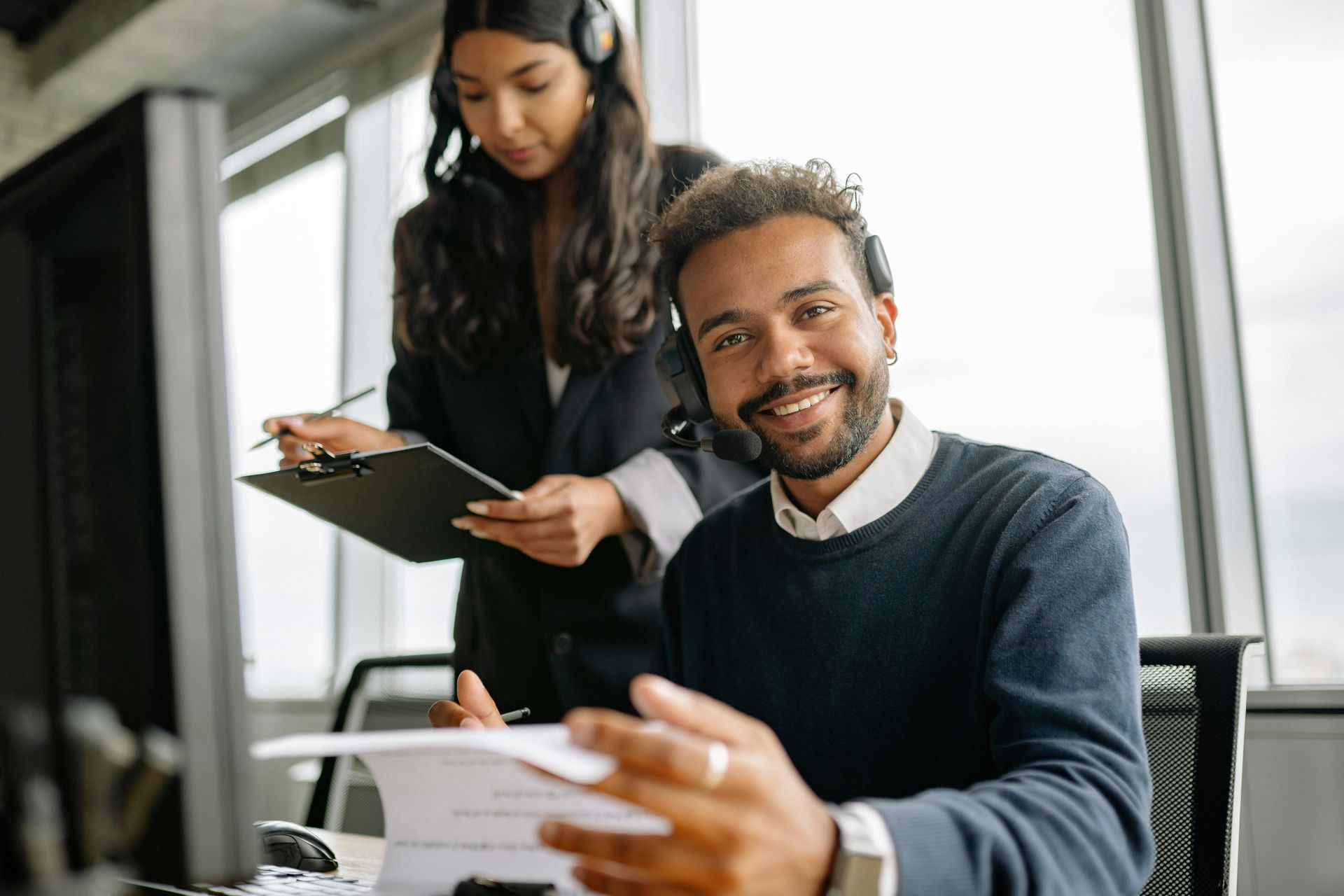 Man smiling at camera while working on a computer, another person looking on, both wearing headsets.