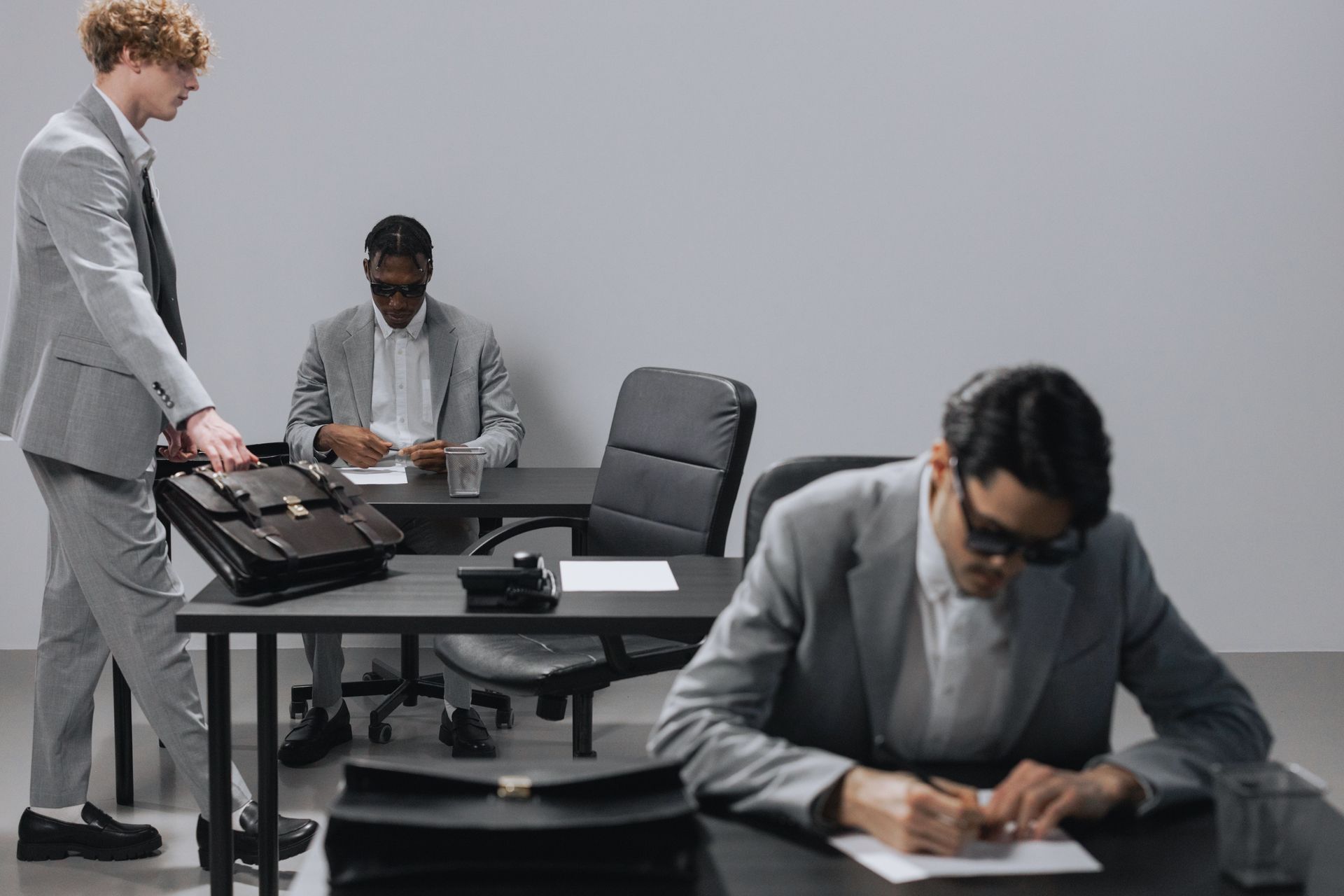 Three people in gray suits in a modern office, one writing in foreground and two seated at a table.