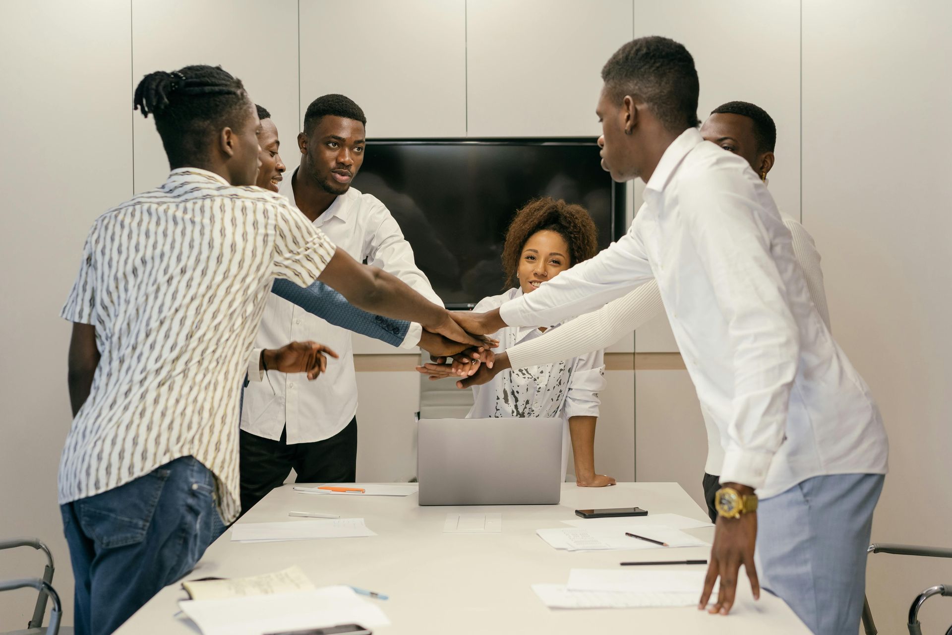 A group of professionals in a modern office gather around a table, placing their hands together in a collaborative gesture.