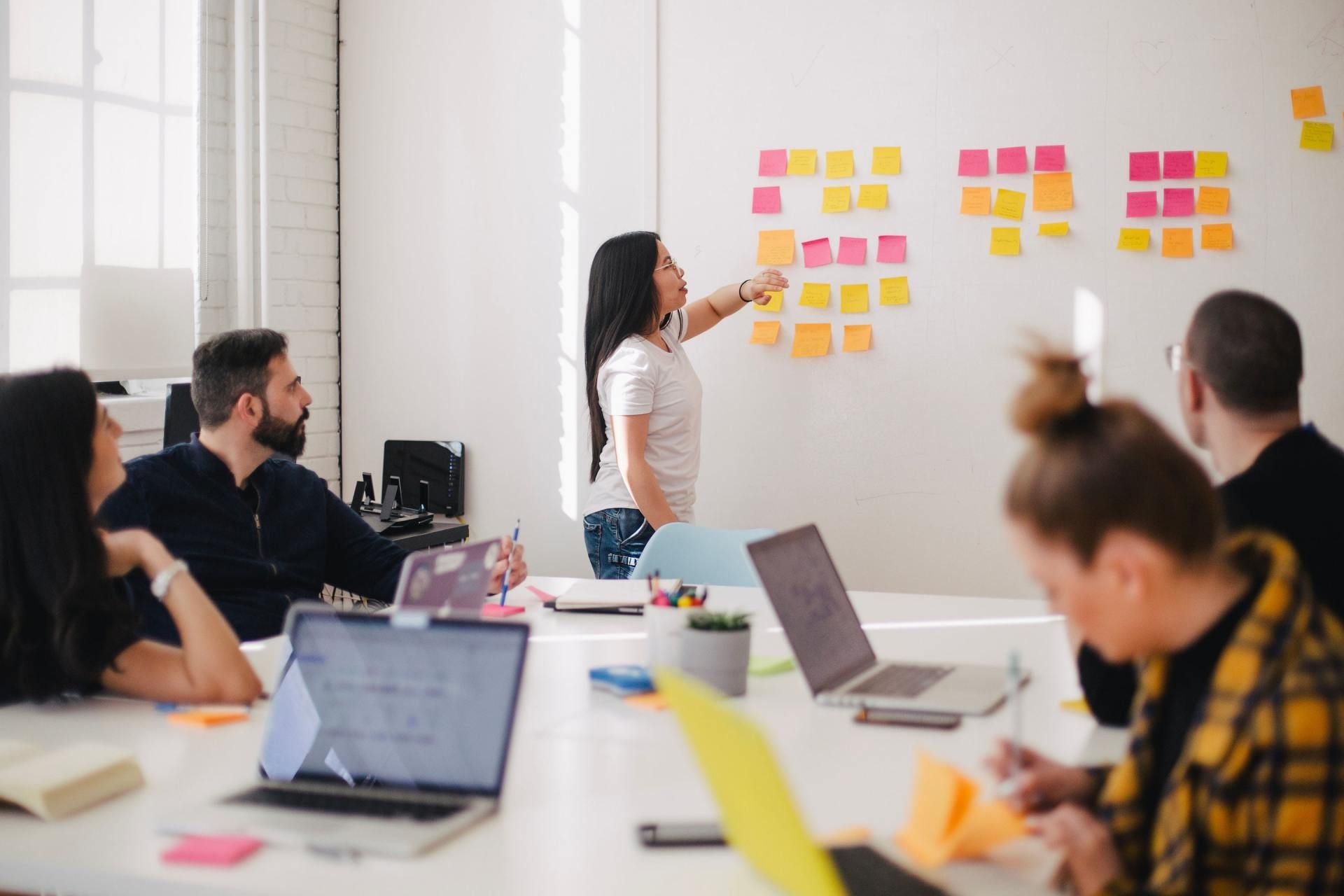 Team meeting around a table. Woman points at sticky notes on whiteboard. Laptops and colorful pens visible.