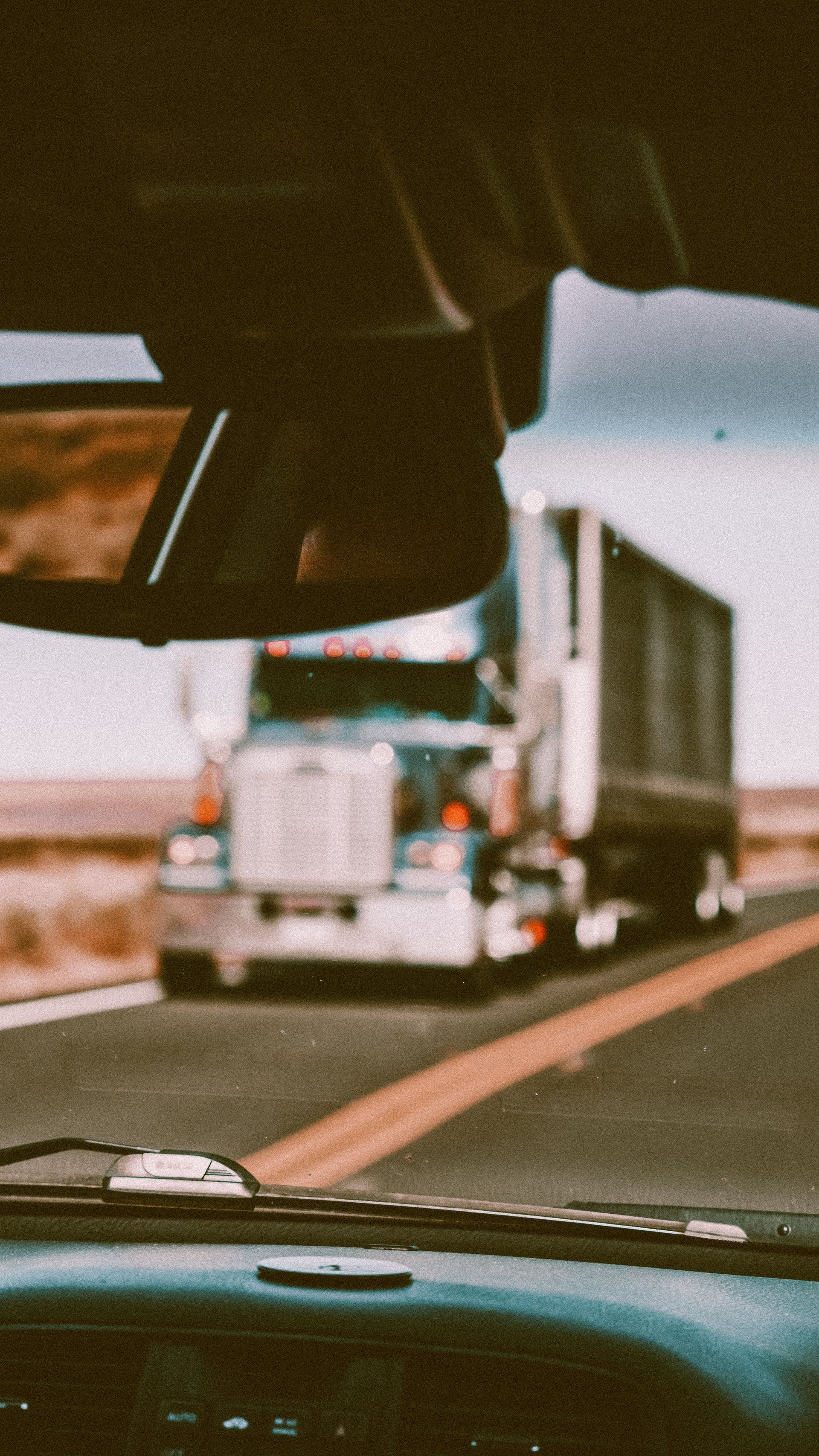 Semi-truck driving on a highway, seen through a vehicle windshield and rearview mirror, overcast sky.