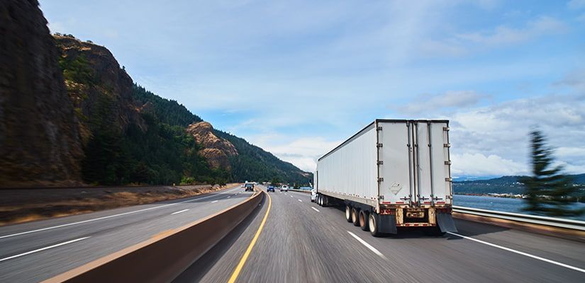 Semi-truck driving on highway with mountains and water in background.