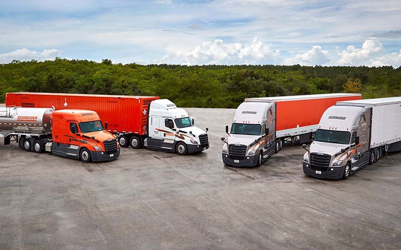 Four semi-trucks parked on concrete; orange, white, and silver in color, with orange and white trailers, set against a tree line.