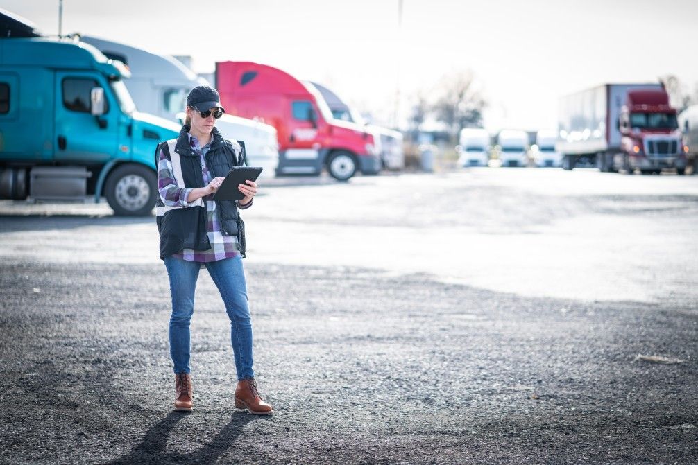 Woman in a cap and vest reviews a clipboard in a truck yard with several semi-trucks parked.