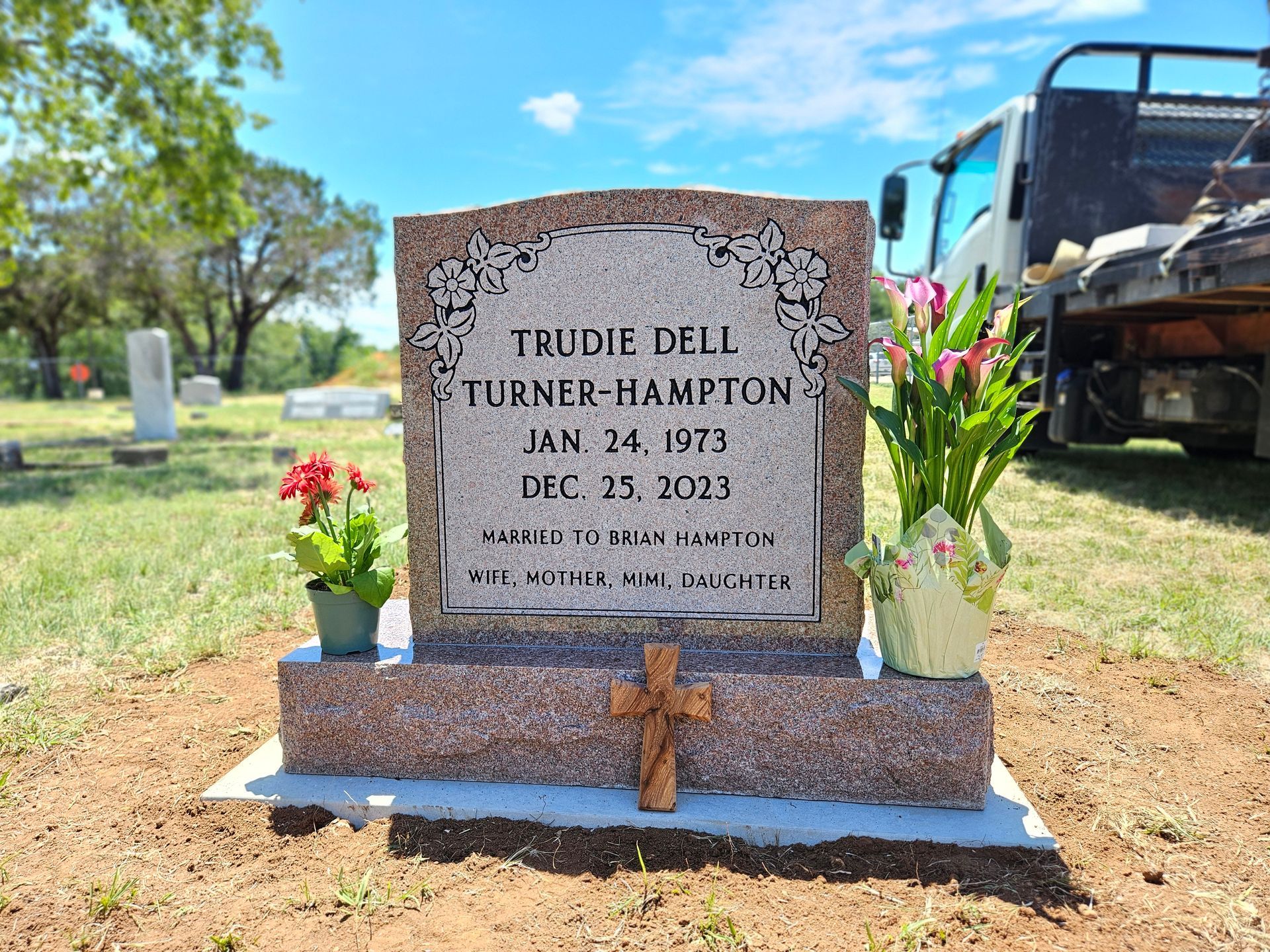 A gravestone in a cemetery with flowers and a truck in the background.