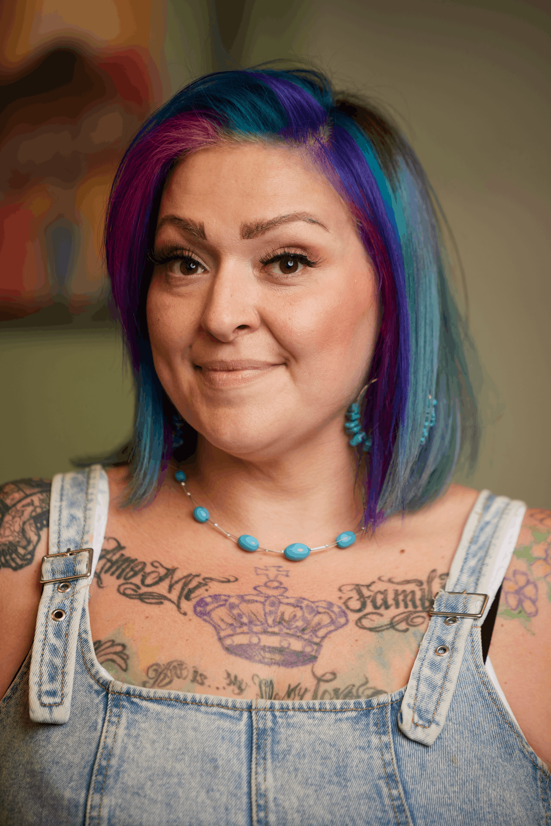 A person with shoulder-length, multi-colored hair poses for a selfie, showing tattoos on their arm and chest.