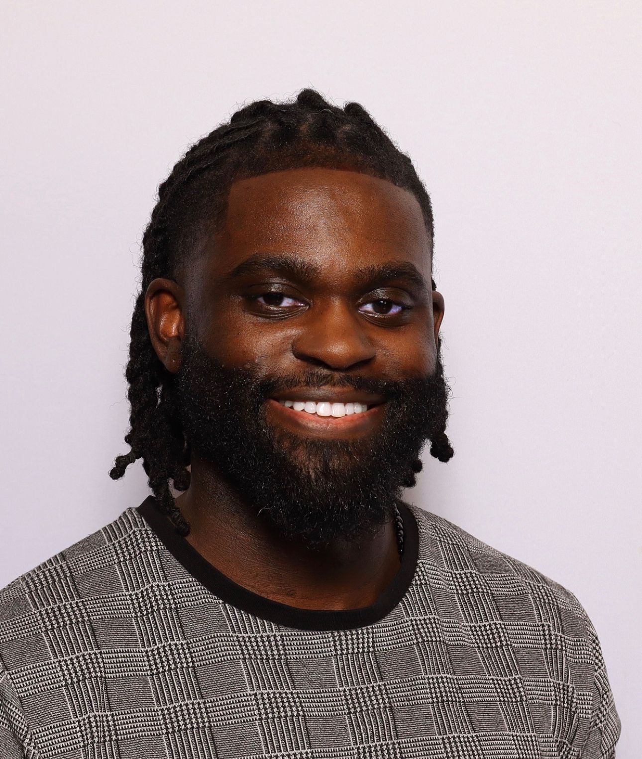A man with a braided hairstyle and a dark beard smiles while wearing a patterned gray sweater against a plain background.